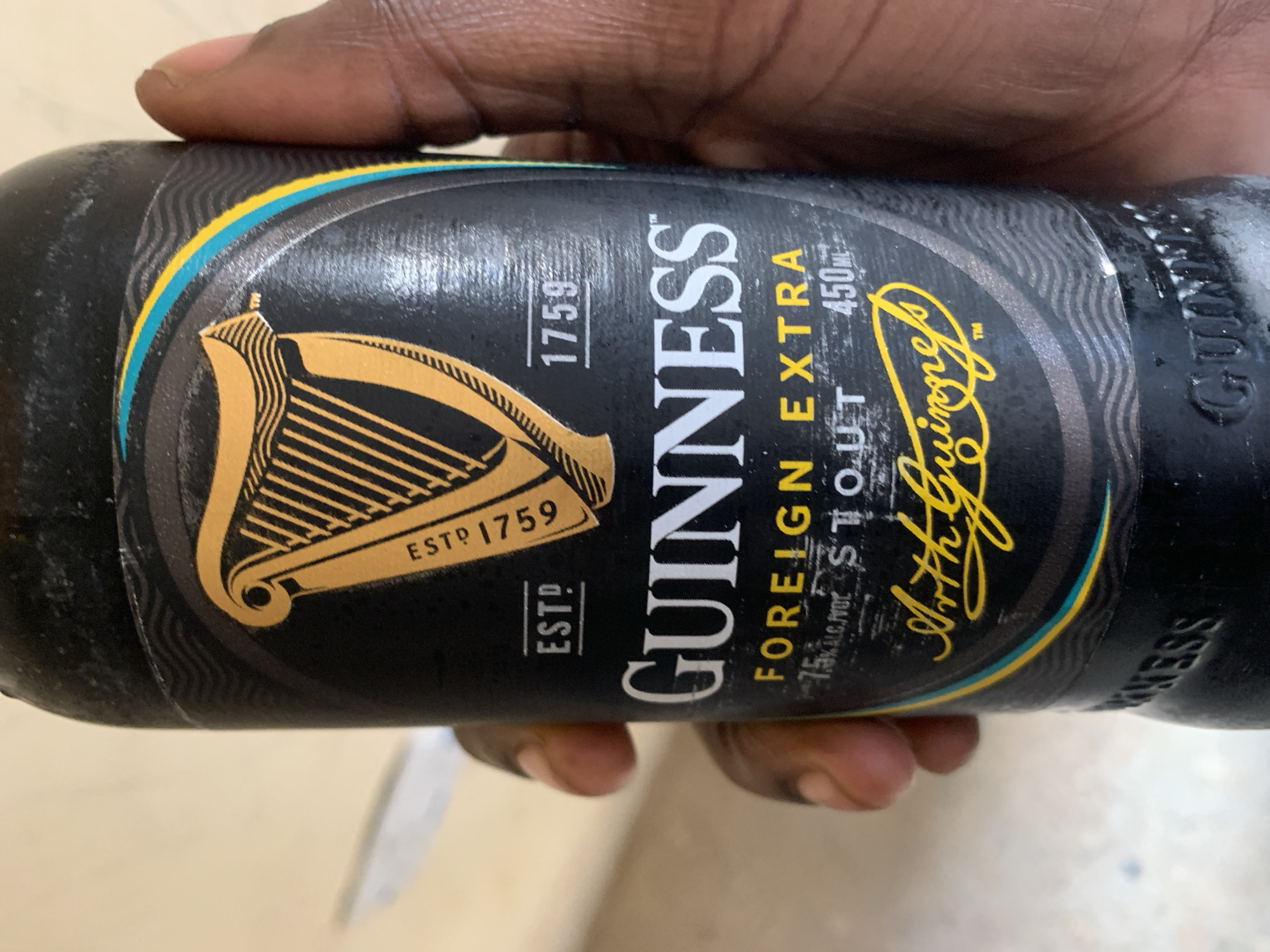A person's hand holds a bottle of Guinness Foreign Extra Stout. The bottle has a black label with the signature Guinness harp logo in gold. The words "Guinness Foreign Extra Stout" are displayed on the label, along with "ESTD 1759" and "450ml". The background is a light-colored surface. There is no indication of a location in the city of New Stadium, Nigeria.