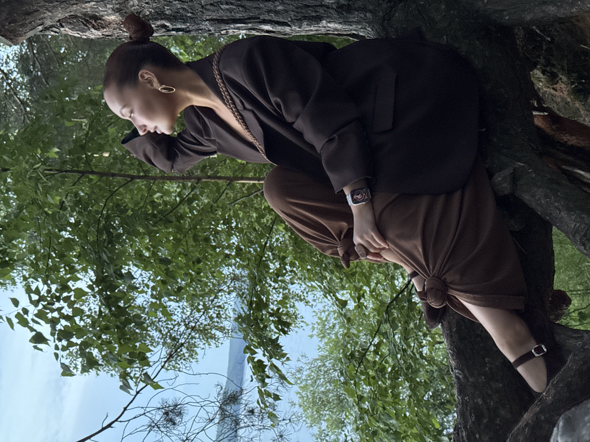 A young woman is pictured in a tree in an outdoor setting. She is wearing a dark brown blazer over a chain detail top, brown pants that are tied at the knee, and dark brown heels with a silver buckle. She has her hair pulled up in a bun and is wearing oval-shaped gold earrings. Her left hand is touching her forehead, and she is looking downwards. A smartwatch is visible on her left wrist. The background consists of green foliage and a bright sky, suggesting a daytime scene. The image is taken from a low angle looking up into the tree. The setting appears to be a natural environment with large tree trunks and branches. No visible text is present in the image.