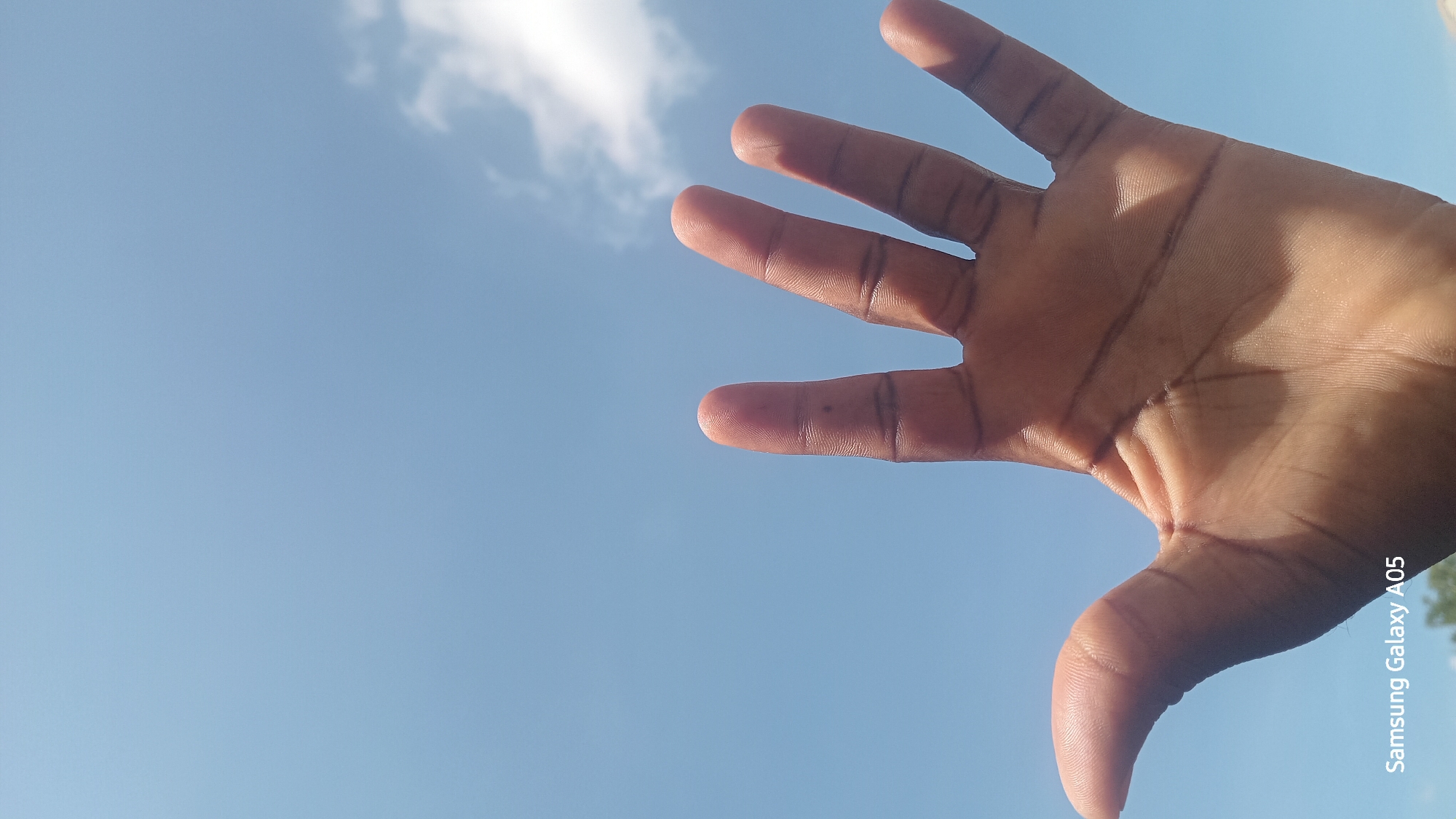 A hand with five fingers outstretched is positioned in the foreground of the image, obscuring the lower portion of a clear blue sky. The sky is dotted with a few white clouds. A watermark reading "Samsung Galaxy A05" is present in the bottom left corner of the image. The image was captured in Ribina East, Nigeria.
