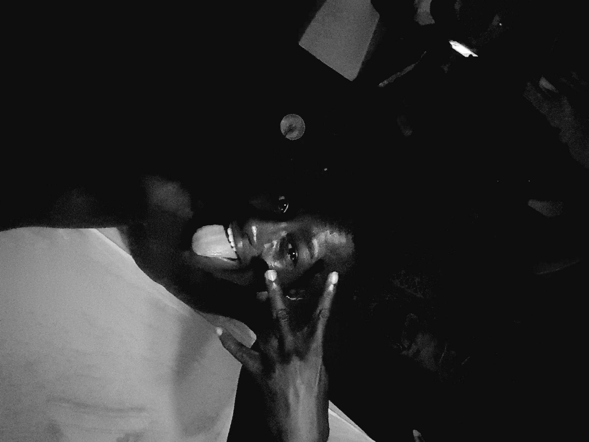 In a dimly lit indoor setting, a person with dark skin and curly hair is captured in a close-up shot. The person's eyes are open, and their tongue is sticking out. Their right hand is raised, with two fingers extended in a peace sign, positioned in front of their forehead. The background is dark and indistinct, with some lighter, blurred shapes suggesting the presence of other people or objects. The overall image has a high contrast, black and white aesthetic. There is no information to indicate the location in Narayi, Nigeria.