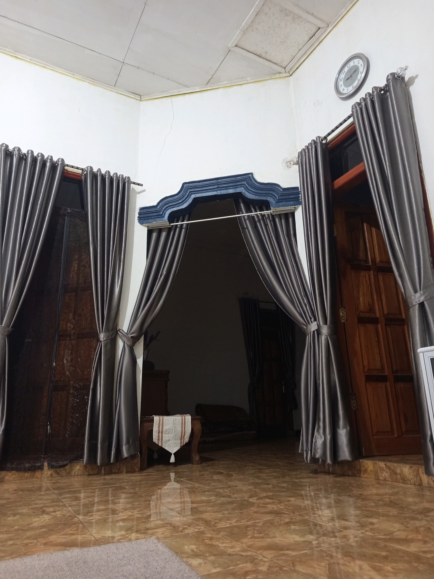 The image depicts an interior view of a room in Murni Jaya, Indonesia. The scene is dimly lit, with a tiled floor reflecting the ambient light. Large, dark gray curtains are drawn across two doorways, one on the left and one on the right. The doorway on the left appears to lead to an outdoor space, as indicated by a dark wooden door. The doorway on the right also features a dark wooden door, elaborately carved. In the center of the image, a smaller opening is revealed, framed by a decorative blue architectural element, leading into a darker interior space. Within this central opening, a small wooden table with a white runner featuring red stripes is visible. A clock with a white face and black hands is mounted on the white wall above the right doorway. The ceiling is a textured white material, with visible seams. The overall impression is of a residential interior, possibly a living room or entryway.