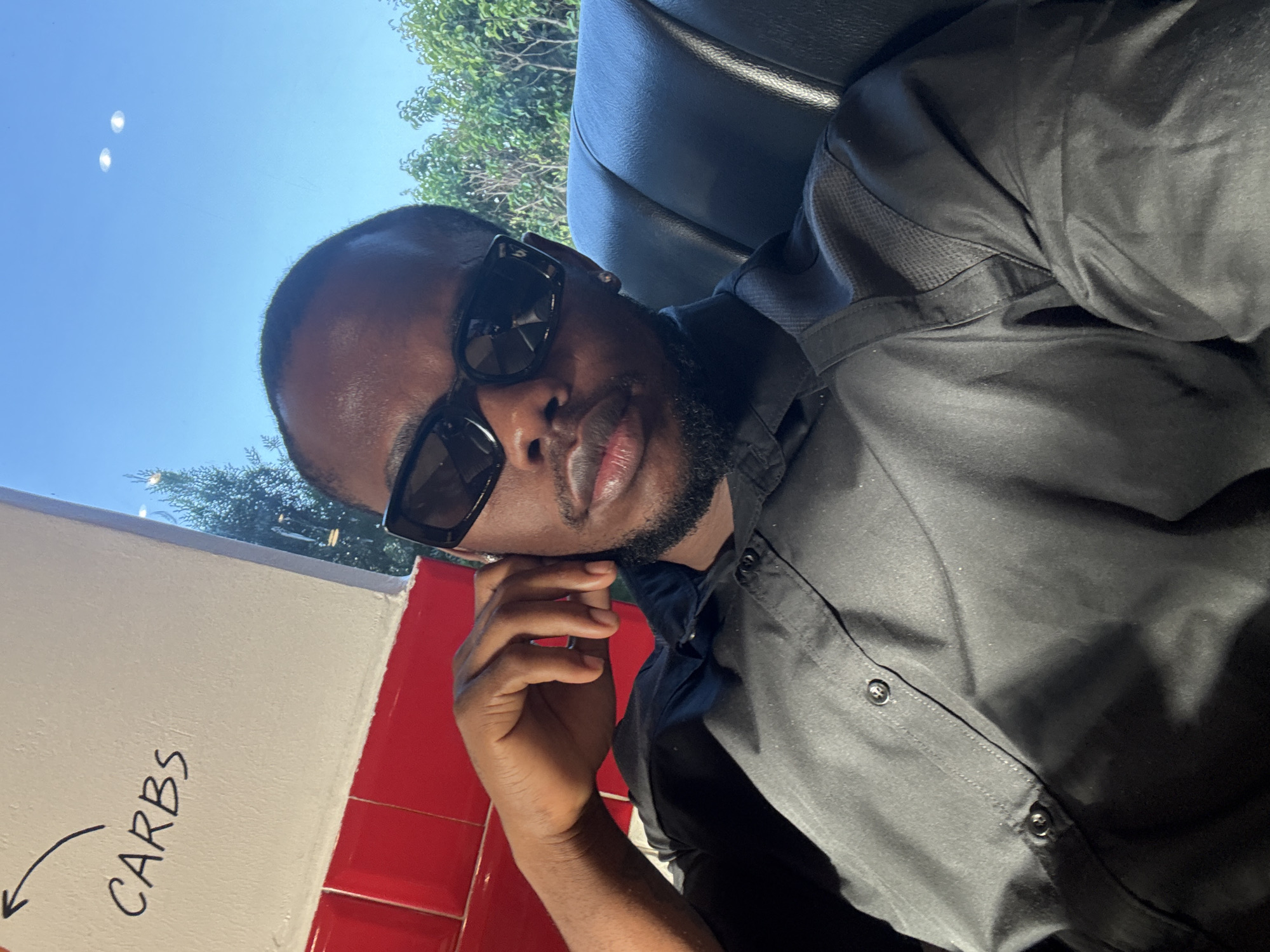 A man with dark skin and a close-cut haircut is sitting indoors wearing dark sunglasses and a black button-down shirt. He has a goatee. His left hand is positioned near his face. Next to him is a white board with the word "CARBS" written on it. The surrounding environment appears to be a restaurant with red tiling. Outside the window, trees and a blue sky are visible. It is not possible to determine the location of Abuja, Nigeria, from the visual information in the image.