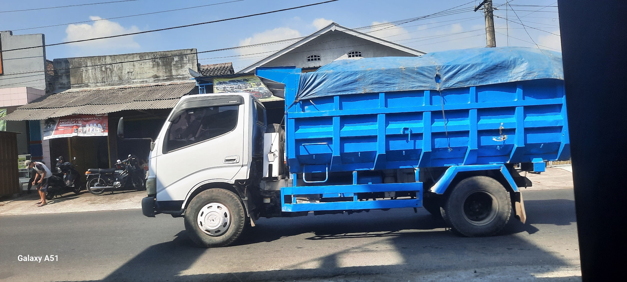 The image depicts a blue tarp-covered truck with a white cab parked on the side of a road in Selopuro, Indonesia. The truck is in the foreground, angled towards the right of the frame. Behind the truck, there are buildings with corrugated roofs and signage. One sign reads "HIGH PERFORMANCE MOTOR OIL". There are several motorcycles parked in front of one of the buildings. A person is visible to the left of the truck, appearing to be adjusting something on a motorcycle. The sky above is blue with scattered clouds, and power lines are visible overhead. The scene appears to be in a commercial or semi-urban area.