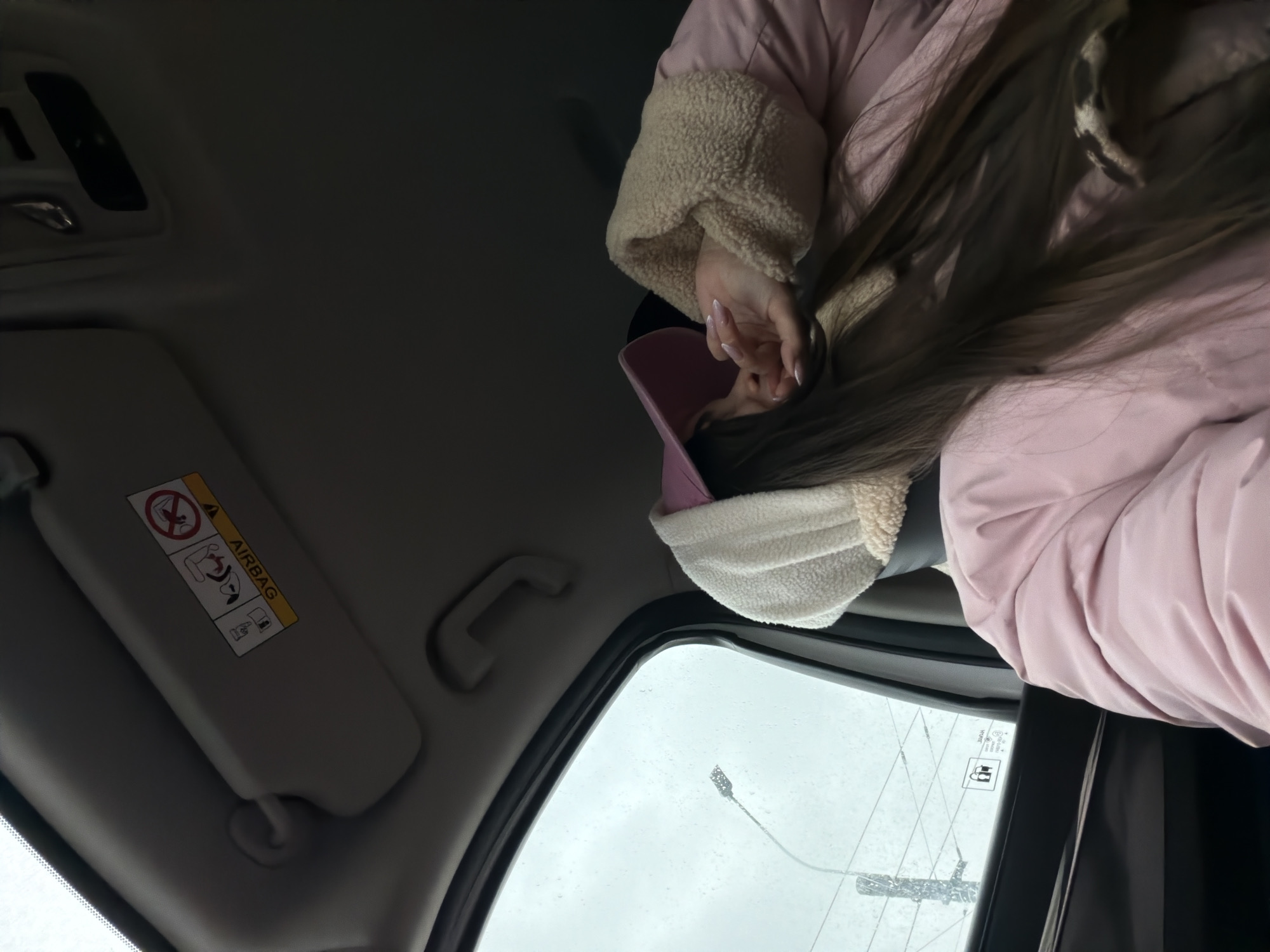 The image shows a view from inside a car. The person in the car is wearing a pink jacket with a sherpa lining and a pink baseball cap. The car's interior is visible, including the sun visor with airbag warnings. Outside the car, there is a streetlamp and overcast sky, suggesting it might be raining. There are also electrical lines and a power pole.
It is impossible to discern if this is Saint Petersburg Russia without further information. The scenery and car interior do not indicate a location.