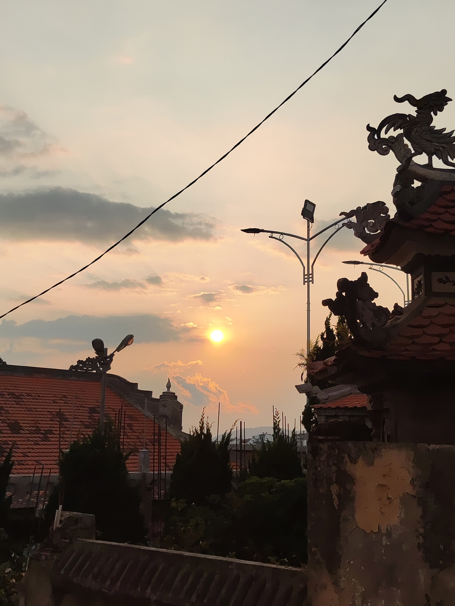 An exterior shot captures a sunset view over Da Lat, Vietnam. In the foreground, there's an aging stone structure, its surface mottled with peeling paint. A traditional Vietnamese building with a terracotta-tiled roof is prominent, adorned with dragon or phoenix figurines at its corners. Further back, more terracotta roofs and spires hint at a dense urban environment. Lush greenery, including cypress-like trees, fills spaces between buildings. Modern streetlights interrupt the scene, contrasting with the traditional architecture. The sky displays a warm sunset palette, with the sun partially obscured by scattered clouds. A power line stretches across the frame.