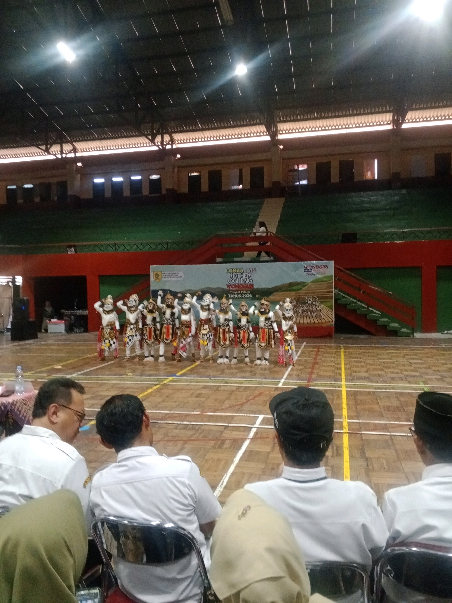 The image depicts an indoor sports hall, likely in Wuryorejo, Indonesia, during an event. In the background, a group of performers in traditional costumes, characterized by white masks and elaborate headdresses, stand in front of a banner. The banner appears to announce a "Lomba Lari" (Running Race) and mentions "Wonogiri" and "Tingkat Pelajar Tahun 2026". The hall has stadium seating in the upper sections and a wooden floor in the main area, marked with court lines. In the foreground, several individuals are seated, viewed from behind, some wearing white shirts. One person wears a black cap, and another wears a peci, a traditional cap. The lighting suggests an indoor event, possibly in the evening or under artificial lights. The overall scene suggests a cultural or sporting competition.