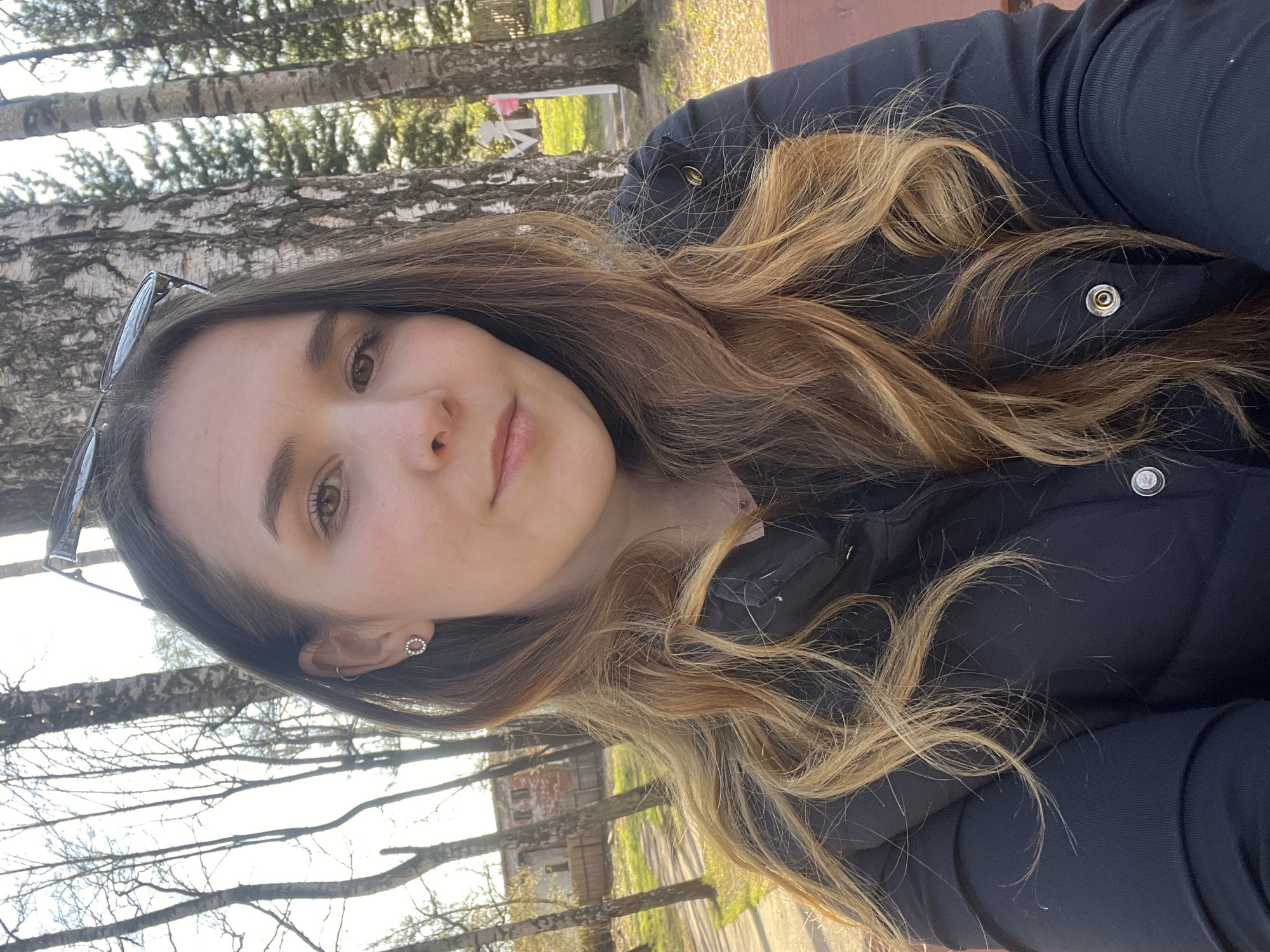 A selfie of a young woman with long brown and blonde hair wearing a dark jacket. She is outdoors in what appears to be a park setting, with birch trees visible in the background. The exact location within the city of Votkinsk, Russia, is not discernable from the image.
