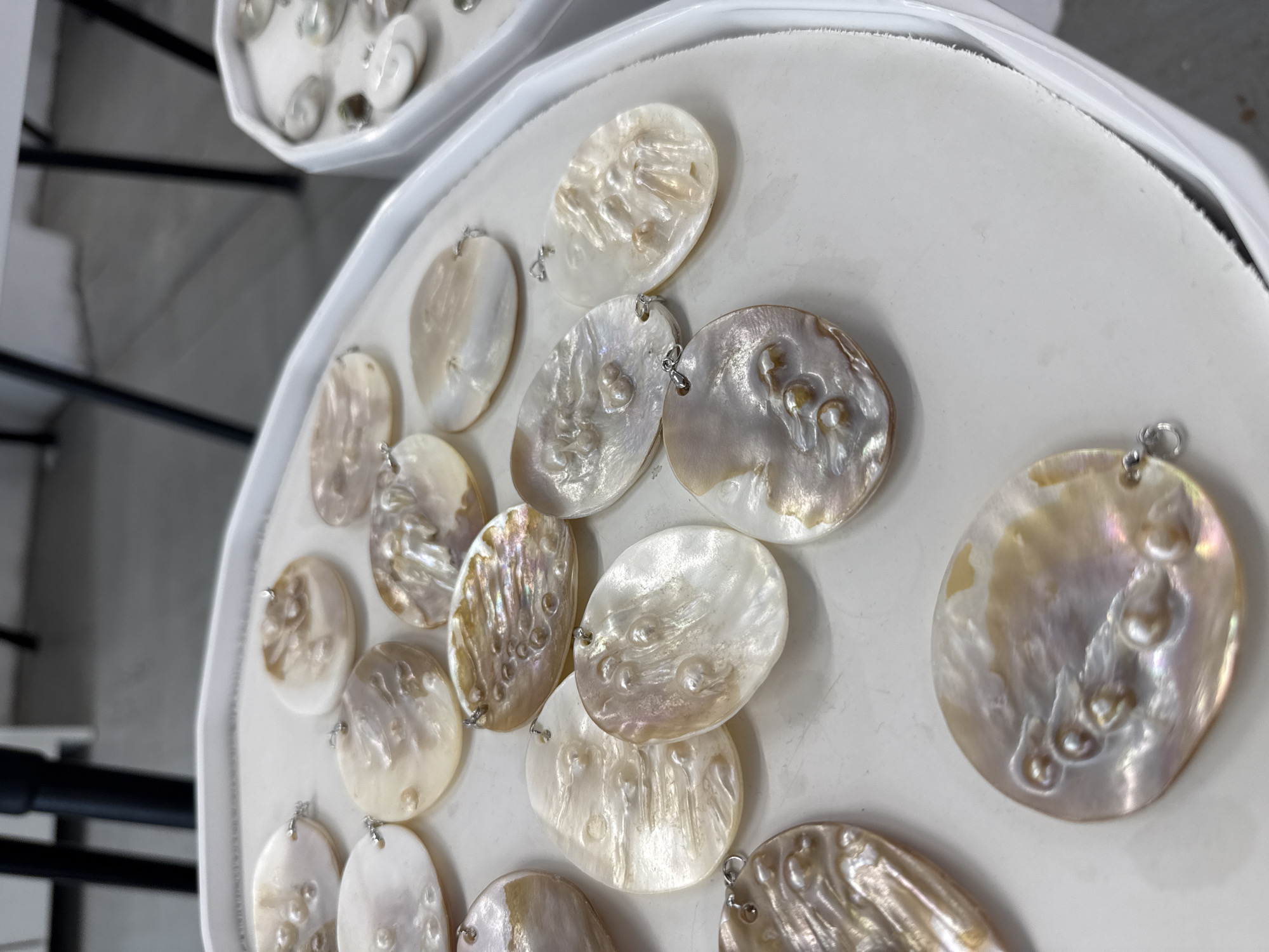 The image shows a close-up view of a white, circular tray filled with oval-shaped pendants made of a pearlescent material, possibly mother-of-pearl. Some pendants have embedded pearl-like embellishments. Each pendant has a small silver-colored loop attached at the top. A second tray of similar pendants is visible in the background. The image was taken in the city of Синьково, Russia.