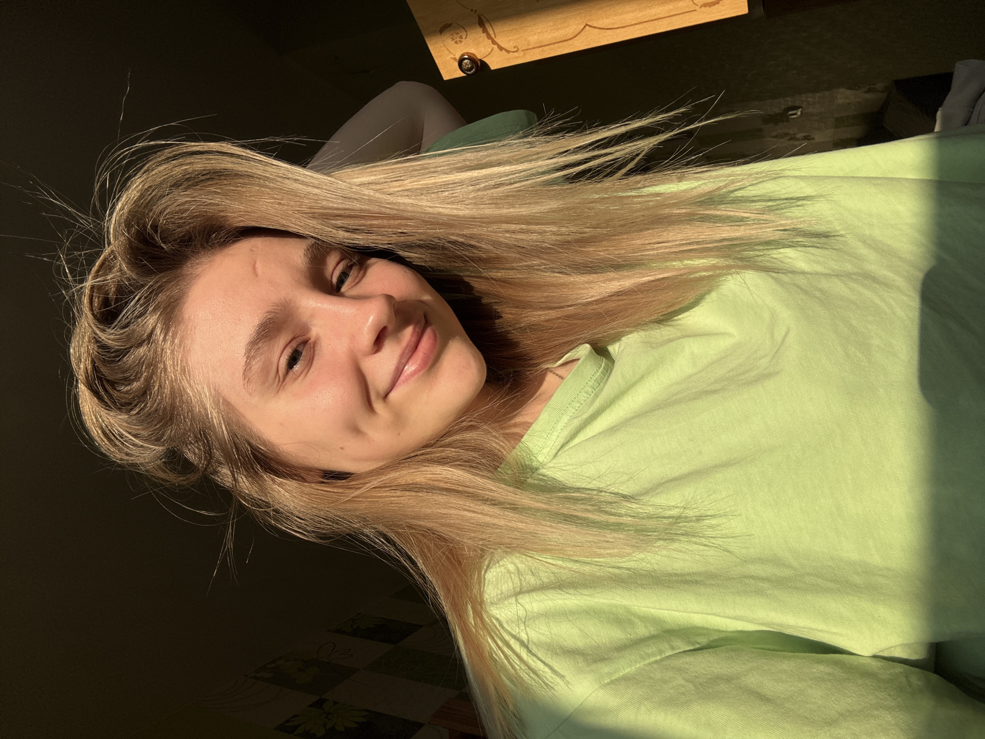 A single female individual is depicted indoors. The subject, appearing as a young adult with long, light-colored hair, is positioned centrally and looks directly at the viewer with a subtle, closed-mouth smile, exhibiting a dimple on her left cheek. She wears a light green, crew-neck, long-sleeved t-shirt. Strong, warm, directional light, consistent with natural sunlight, illuminates the right side of her face and hair, casting shadows on her left side and the background. The background consists of an interior space. On the left, a dark wall displays a faintly patterned wallpaper, possibly with a grid or floral design. On the right, a section of a light-colored wooden structure, potentially a piece of furniture like a cabinet door, is partially visible, featuring a circular knob or decorative element. The precise location within the city of Izhevsk, Russia, cannot be determined from the visual evidence presented in the image.