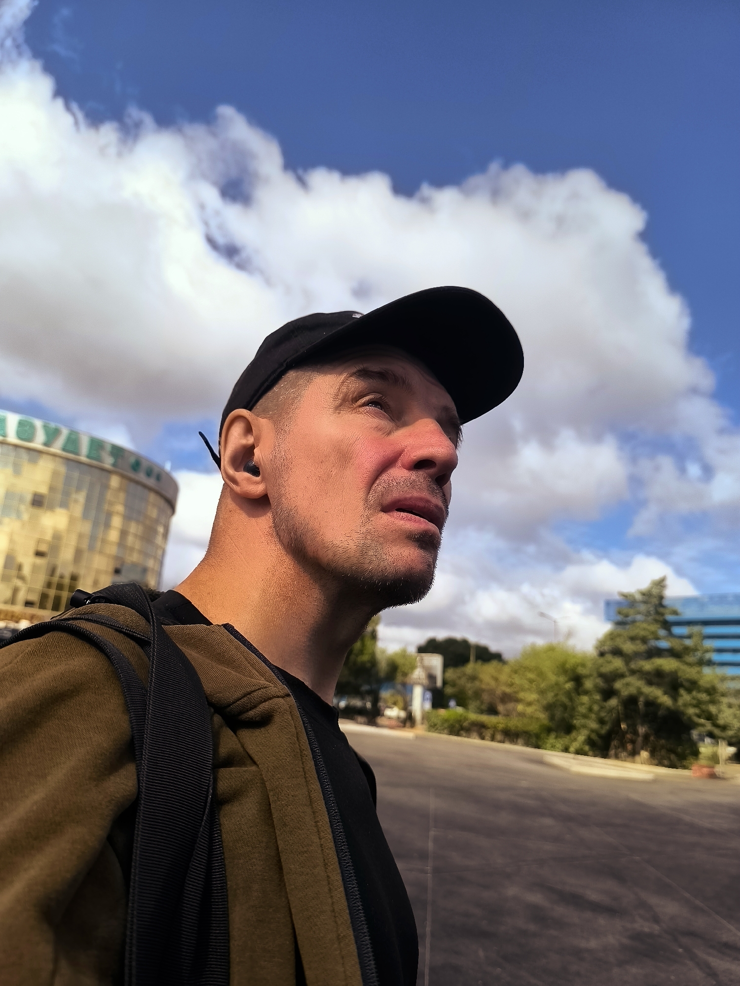 The image shows a man wearing a black baseball cap, a brown jacket over a black t-shirt, and a backpack. He has an earbud in his left ear and is looking up and to his right. The background features a building with a gold-colored facade, a street, trees, and a bright blue sky with white clouds. The building in the background has Cyrillic lettering that reads "ҚАРАҒАНДЫ ҚАЛАҺЫ ӘКІМДІГІ," which translates to "Qarağandı City Administration." The location is Qarağandı, Kazakhstan.
