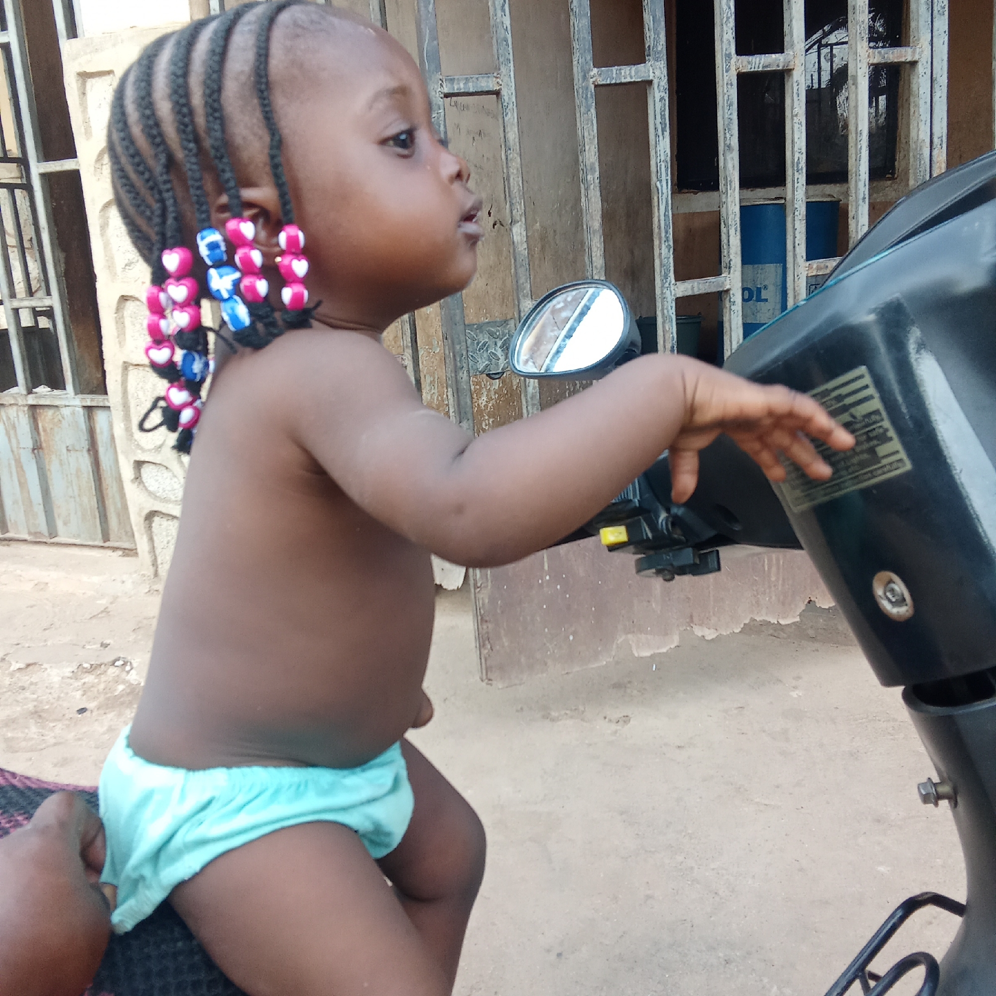 This outdoor photograph features a young, dark-skinned child, likely a toddler, seated on or beside a black scooter or motorcycle. The child's hair is styled in multiple short, dark braids, each decorated with vibrant pink and blue heart-shaped beads. They are wearing light blue, elastic-waisted briefs or shorts. The child is captured in profile, looking intently towards the right with an open-mouthed, curious expression. Their left hand rests gently on the scooter's front panel, near a rectangular sticker with illegible text and a visible side mirror.
The background reveals a rustic, light-colored building with dark metal grates over what appears to be windows or a doorway. Through these grates, a dark interior is visible, along with a blue barrel that bears partially visible text, "DL" and "OL" (possibly "OIL"). The ground beneath the child and scooter is unpaved and dusty. The lighting suggests it is daytime. The scene depicts a casual moment in an outdoor setting, characteristic of Akwanga, Nigeria.