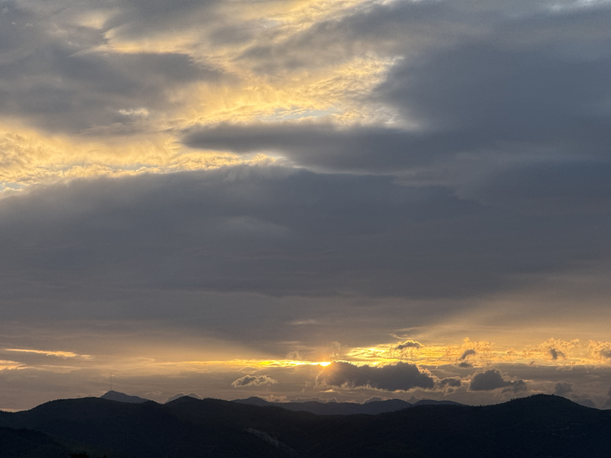 The image captures a sunset over the mountainous landscape of Song Cau, Vietnam. The sky is filled with dramatic clouds, with streaks of golden light piercing through the darker, gray masses. The sun is partially obscured by clouds, casting a warm glow on the horizon. Below the sky, silhouetted against the sunset, are the undulating forms of mountain ranges, their peaks appearing dark and indistinct. A lone, slender antenna-like structure is visible atop one of the distant mountains. The foreground is dominated by the dark shapes of the mountains, suggesting a vantage point from a higher elevation looking out over the landscape.