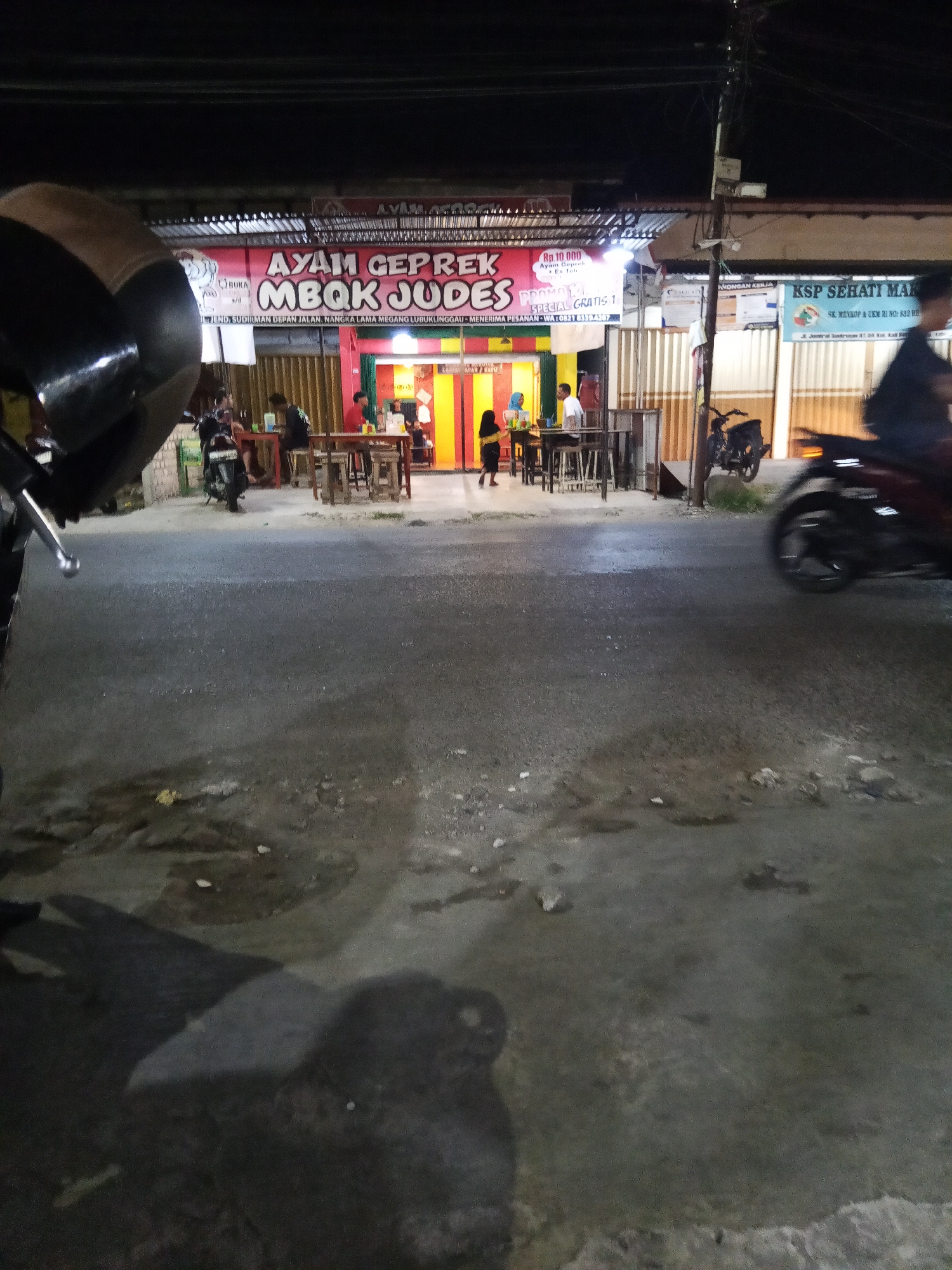 This is a nighttime, eye-level, medium shot of a street in Lubuklinggau, Indonesia, focused on a food establishment. The establishment features a prominent red banner advertising "AYAM GEPREK MBOK JUDES" with prices and contact information. There are tables and chairs set up outside for dining. Several people are visible, some seated at tables and others standing. A motorcycle is parked in front of the establishment, and another motorcycle is seen driving past in the foreground. The street itself is paved and appears wet in places, with some debris scattered on the surface. The lighting suggests it is after dark, with artificial lights illuminating the scene.