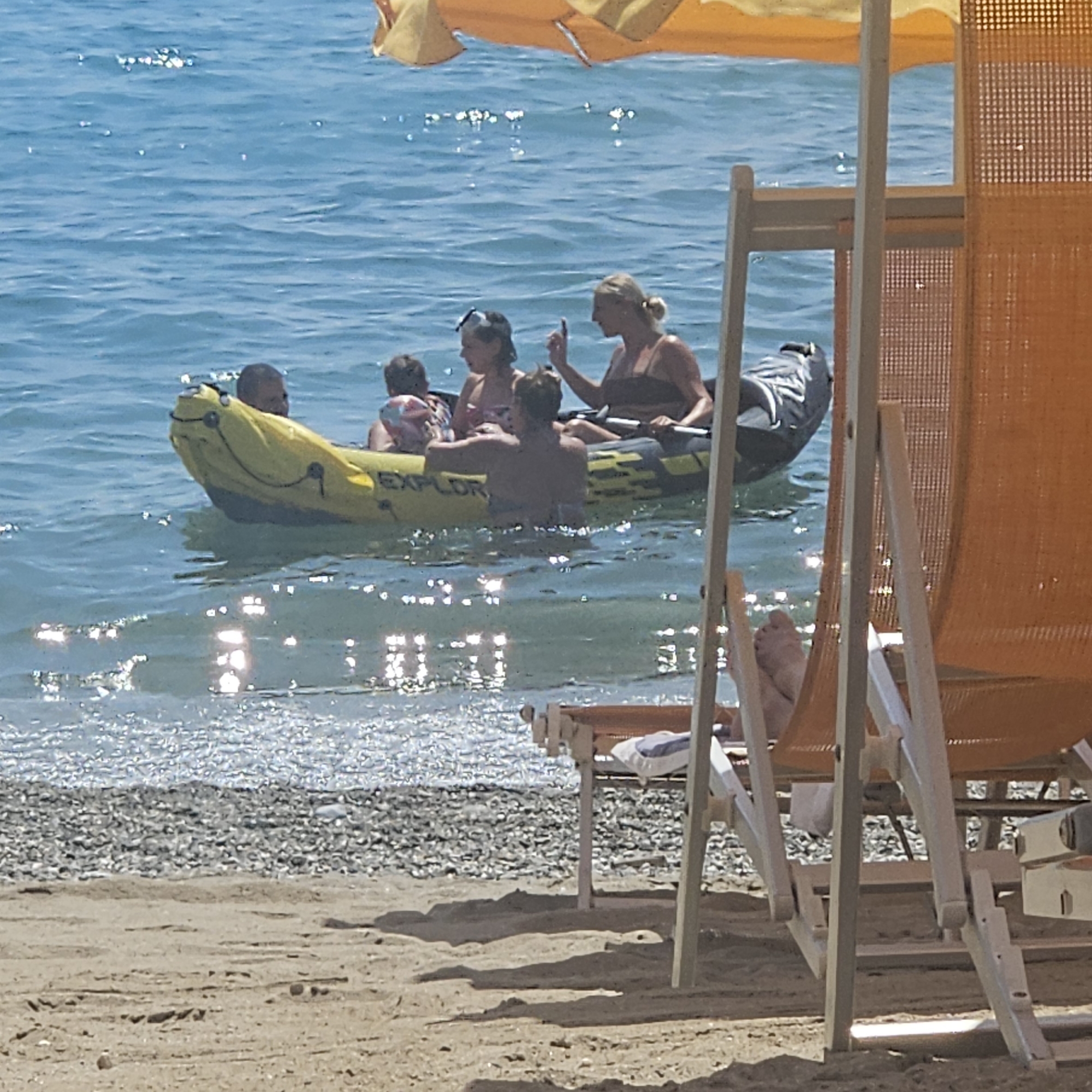 A yellow inflatable raft with the word "EXPLOR" printed on its side is floating in the blue sea. Five people are in the raft. In the foreground, two lounge chairs, one orange and one white, are set up on a sandy beach with a pebble shoreline behind it. The water is calm and reflects the sunlight. An orange umbrella is visible in the upper left corner of the image. The scene appears to be a beach in the city of Savona, Italy.