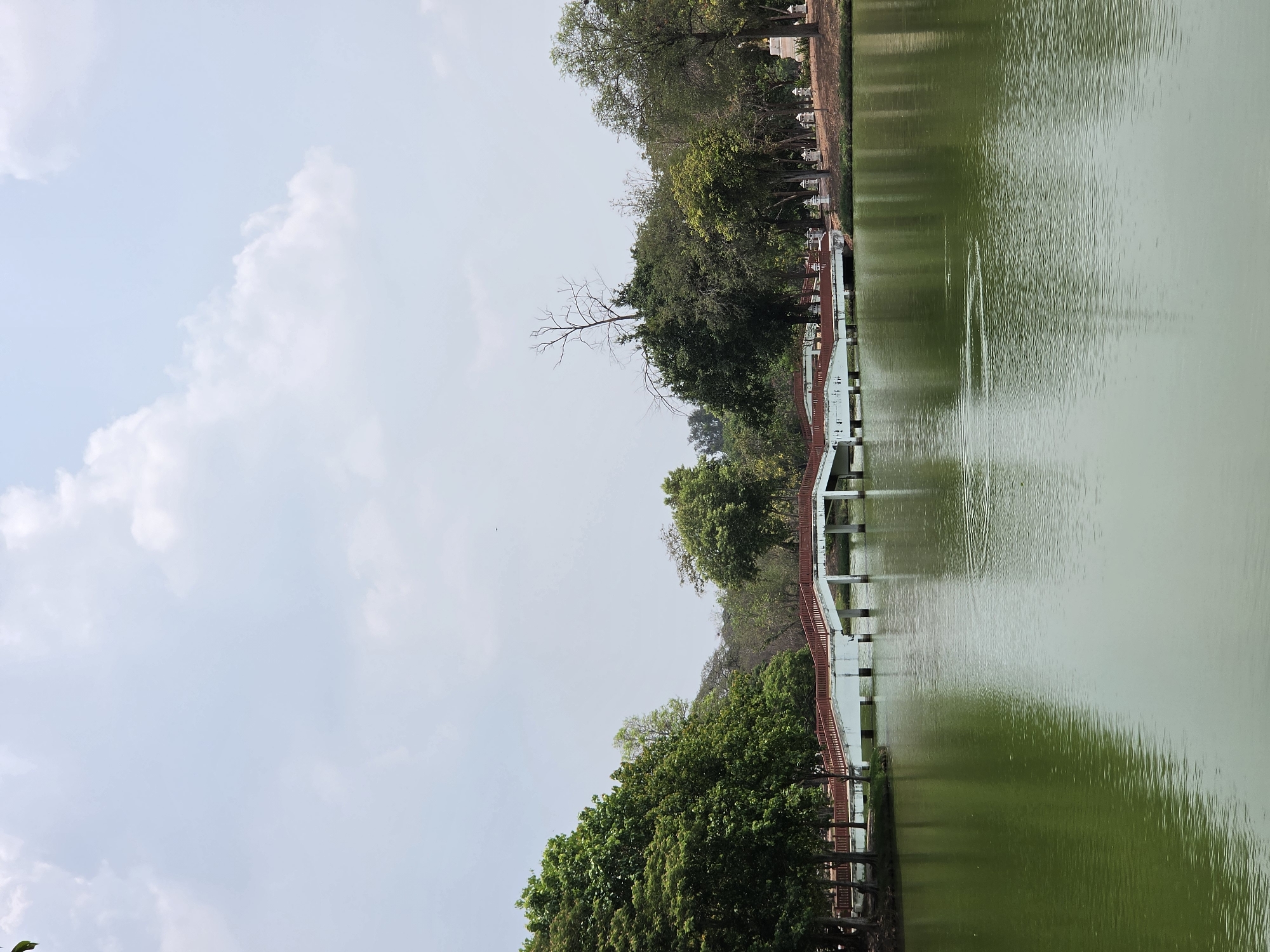 The image depicts a tranquil scene of a lake or pond, likely located in the city of Phra Nakhon Si Ayutthaya City Municipality, Thailand. The water is a green color. A bridge crosses the water, constructed of concrete pillars with red railings along the sides. Trees with green foliage line the banks of the lake. The sky is partially cloudy.