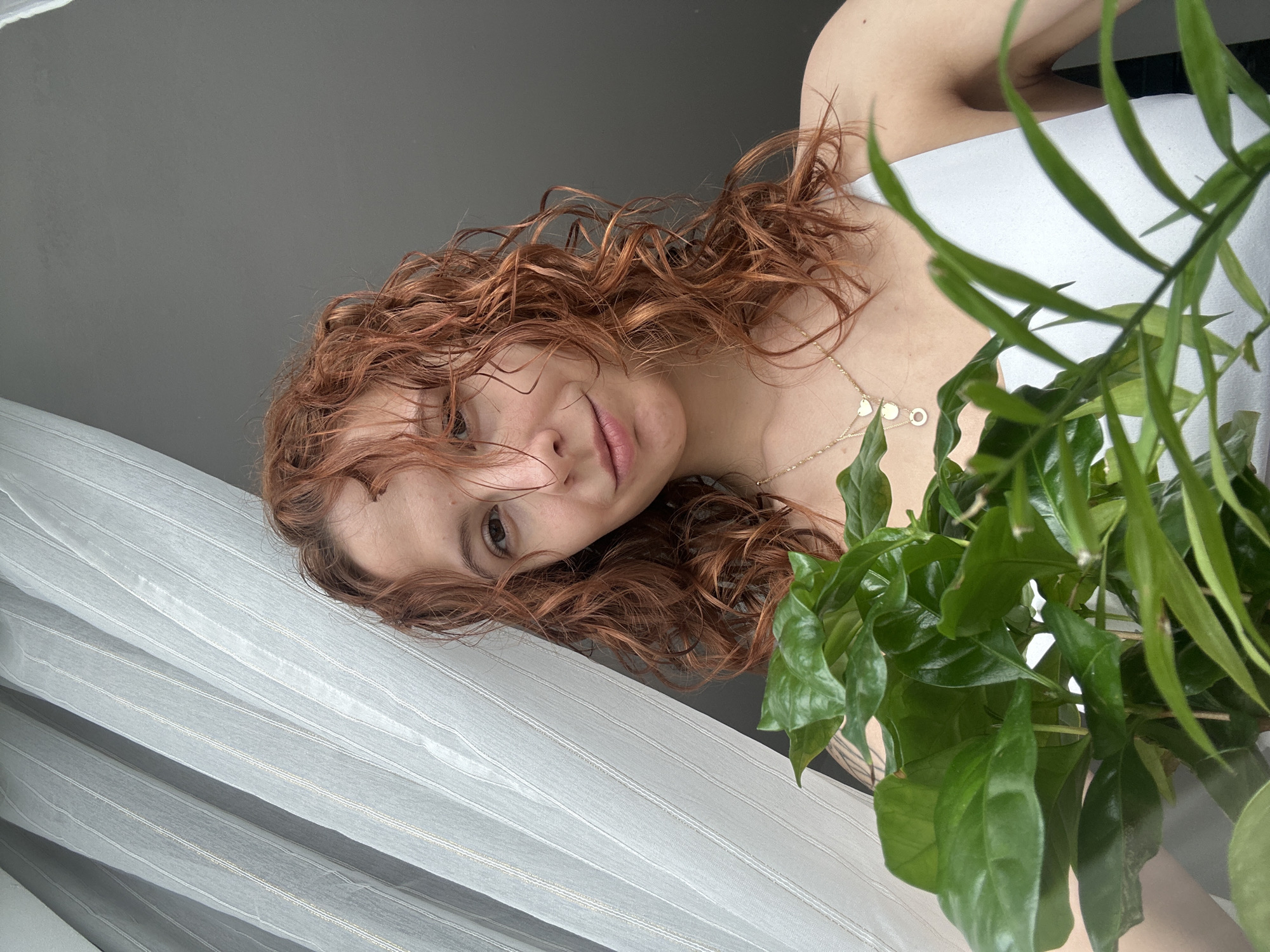 A fair-skinned female with reddish-brown curly hair is positioned on the left side of the image frame. She is wearing a white sleeveless shirt and a gold necklace with circular and heart-shaped pendants. Her gaze is directed towards the camera. To her right, a green leafy plant partially obscures the view. She is standing in front of a window covered with a white, semi-transparent curtain. The background includes a portion of a gray wall or ceiling. The exact location within the city of Novi Sad, Serbia, cannot be determined from the visual information provided.