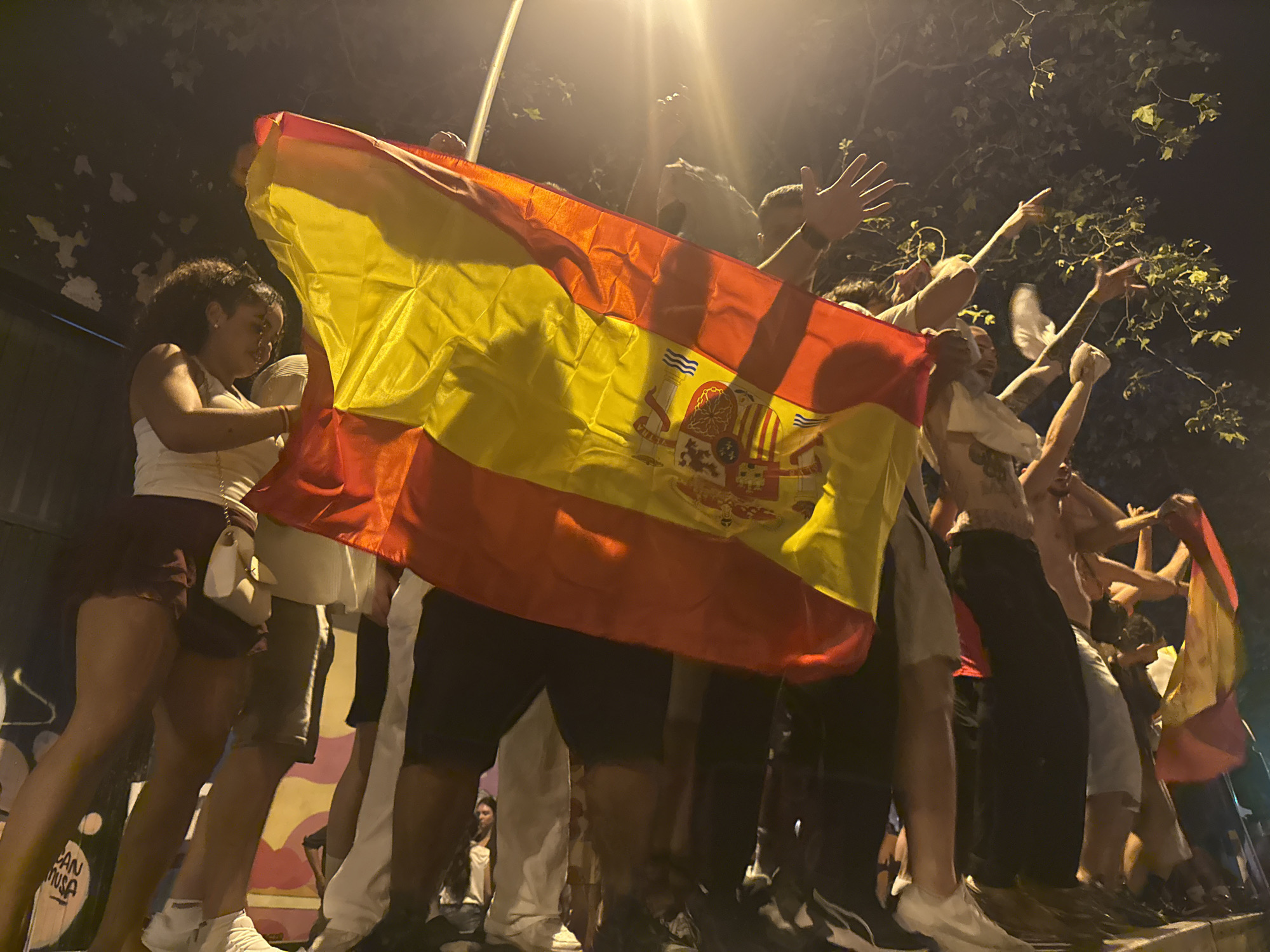 The image shows a group of people holding a large Spanish flag at night in Barcelona, Spain. Many of the individuals are raising their arms in celebration or excitement. The flag, predominantly red and yellow, is spread across the foreground, obscuring parts of the people behind it. In the background, trees are visible, suggesting an outdoor setting. The lighting indicates it is dark, with artificial light sources contributing to the scene's illumination. The people appear to be standing on a raised surface, possibly a wall or a stage. The overall atmosphere seems festive and energetic.