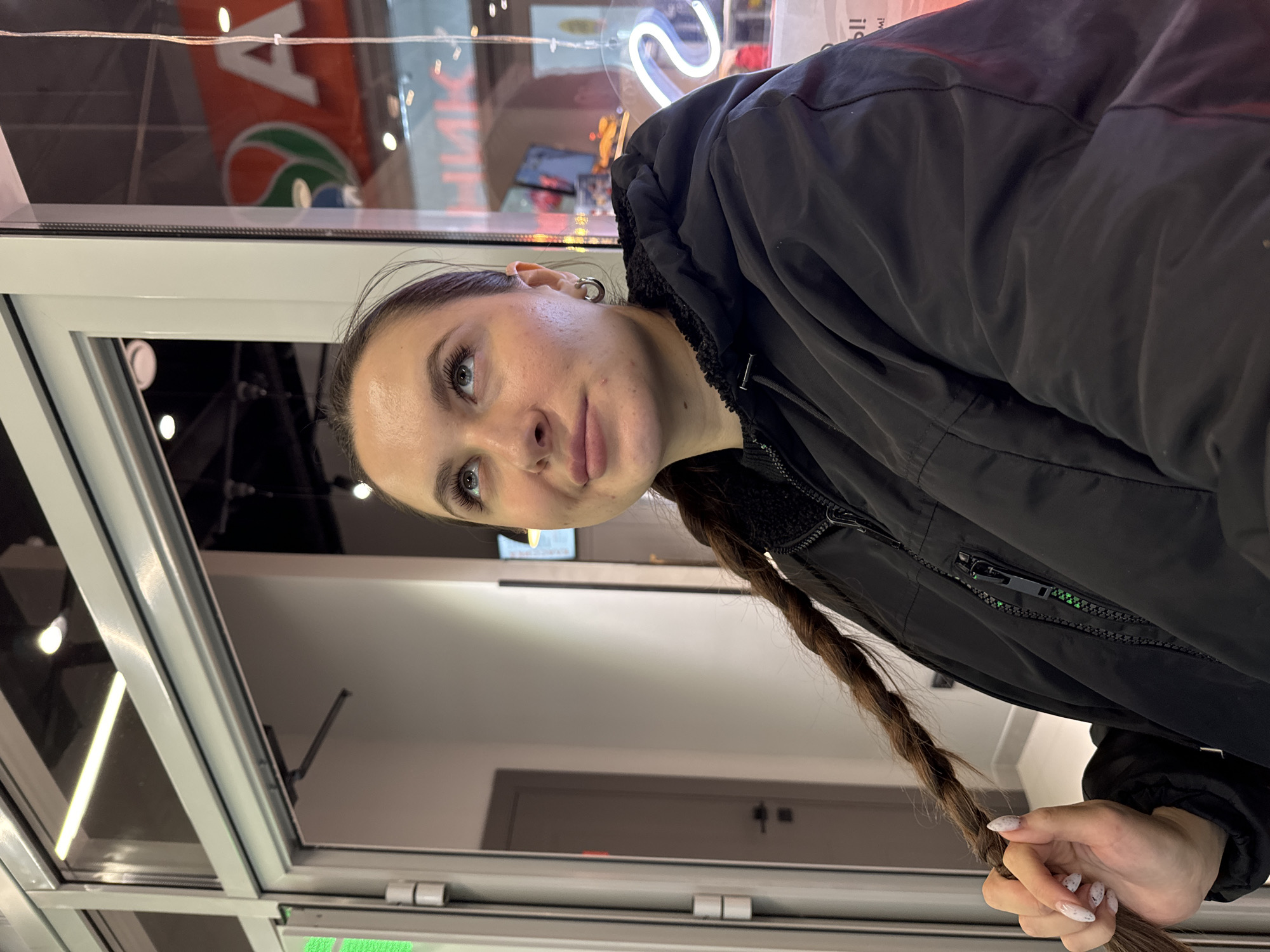 A woman with fair skin and brown hair braided down her shoulder stands in what appears to be a storefront or entrance in Odintsovo, Russia. She wears a black hooded jacket. The location seems to be near a window, which reflects a sign with Cyrillic characters and a neon light. A glimpse inside shows a neutral-colored wall and a door. The woman is holding the end of her braid.