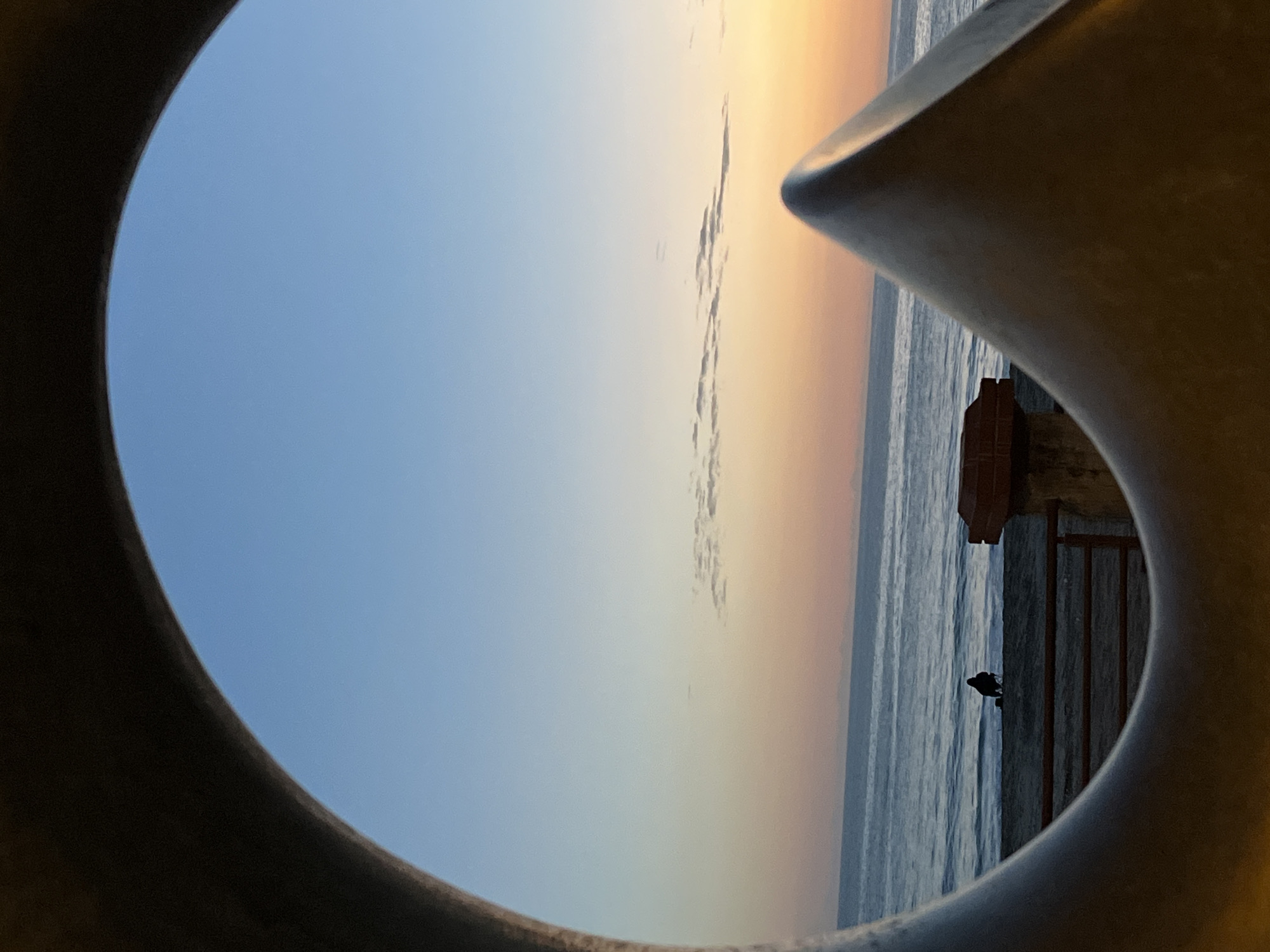 The image shows a view of the sea and sky at sunset through a circular opening, possibly part of a structure. The sky transitions from blue to orange hues, indicating the time of day. A dark object, possibly a bird, is perched on a structure near the sea. The location is likely in Alanya, Turkey, based on the typical coastal scenery of the region. A fragment of a building is visible.