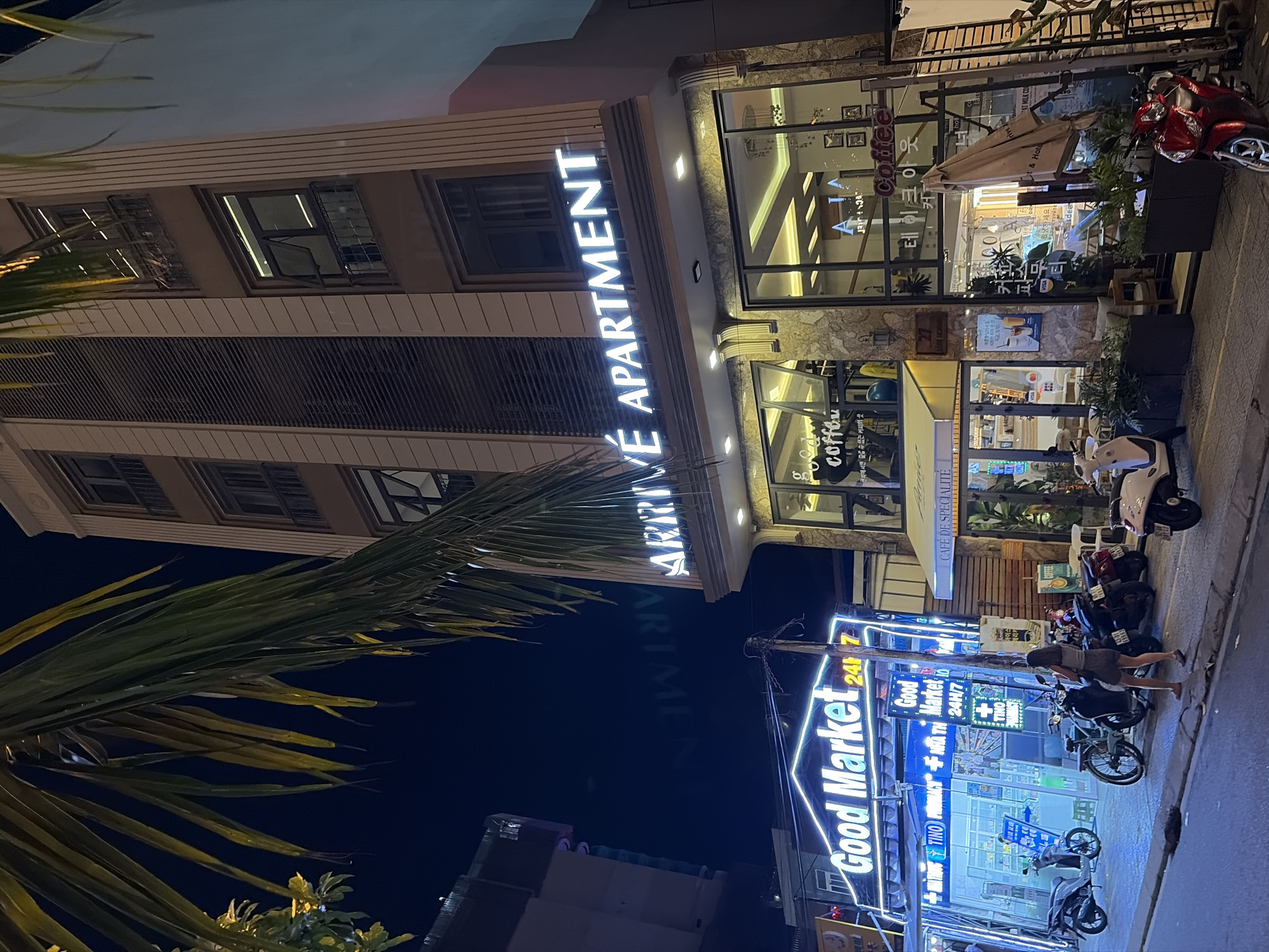 The image captures a street scene at night in Da Nang, Vietnam. In the foreground, a row of buildings lines the street. The building on the right is a multi-story apartment complex with the illuminated sign "ARRIVE APARTMENT" prominently displayed. Below the apartment sign, the ground floor features large glass windows revealing a well-lit interior, possibly a cafe or restaurant, with tables and chairs visible. The signage "coffee" and Korean text are also visible on this building's facade.
To the left of the apartment building, another establishment with bright neon signs reads "Good Market 24H/7". This appears to be a convenience store or market, with shelves stocked with items visible through its windows. Several motorcycles are parked along the street in front of these businesses, indicating common modes of transportation in the area. A lone woman is walking away from the viewer in front of the "Good Market" sign, and another woman is seen through the window of the market. The street is paved, and overhead wires are present. A palm tree with its fronds extending into the frame from the top left adds a tropical element to the urban setting. The overall atmosphere is that of a busy commercial street at night.