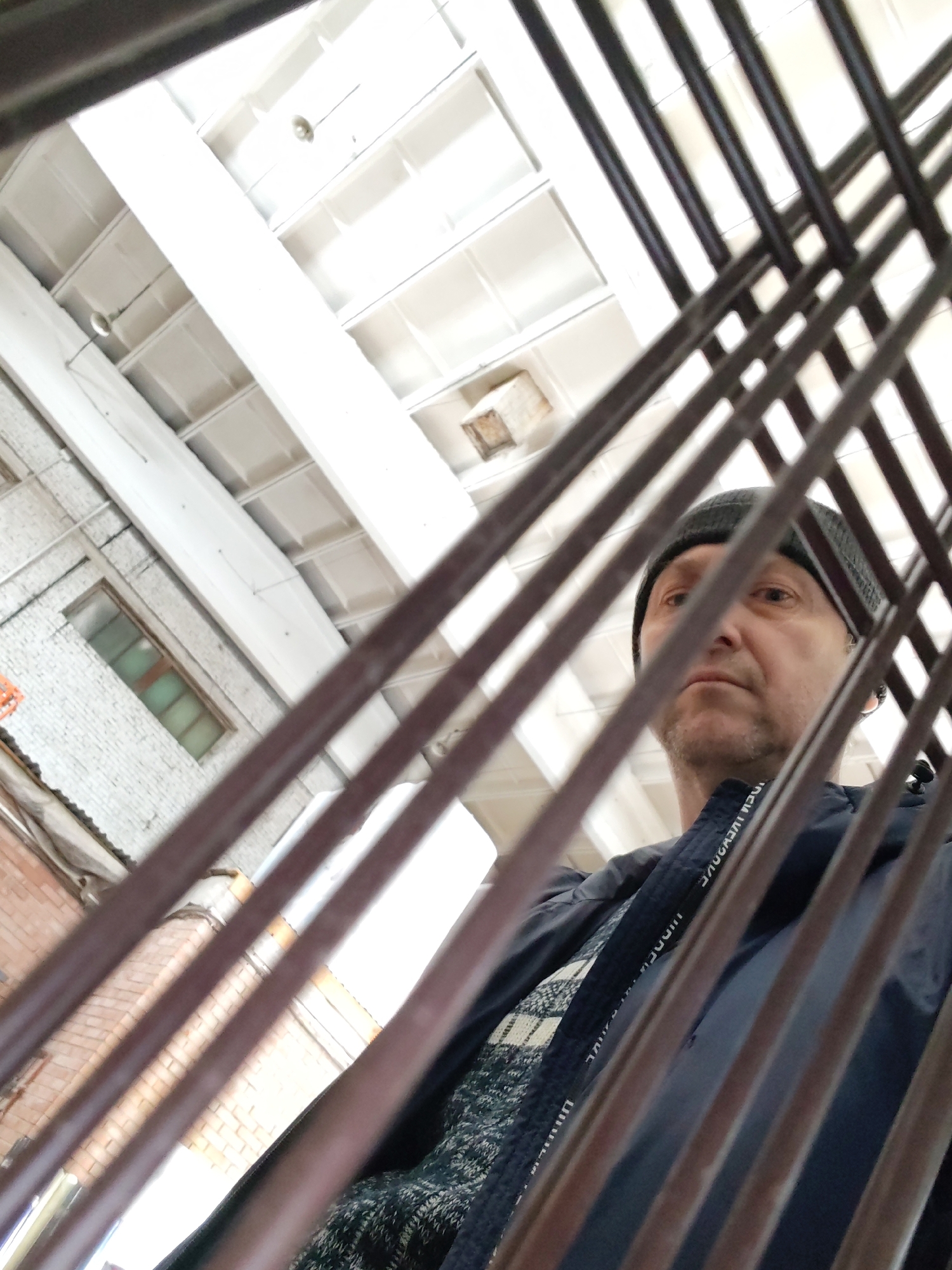 The image shows a man viewed through a prominent metal grid, likely from a low angle looking upwards. The man, wearing a dark beanie and a dark blue jacket over a patterned sweater, is partially obscured by the grid but his face is visible, with a neutral and observant expression as he looks slightly to the right.
The setting is an indoor industrial or warehouse environment, located in Chaikovsky, Russia. The background features a high, white industrial ceiling with exposed beams and recessed panels, fitted with circular light fixtures. To the left, a light-colored, possibly painted, brick wall with a rectangular window and some pipes is visible. Further left, a corrugated metal structure can be discerned.
The metal grid in the foreground creates a strong diagonal and horizontal pattern across the image, contributing to a unique, somewhat obstructed perspective. The overall lighting suggests it is daytime. Text is visible on a strap of the man's jacket, repeating "UNDERWEAR" as part of a design or brand.