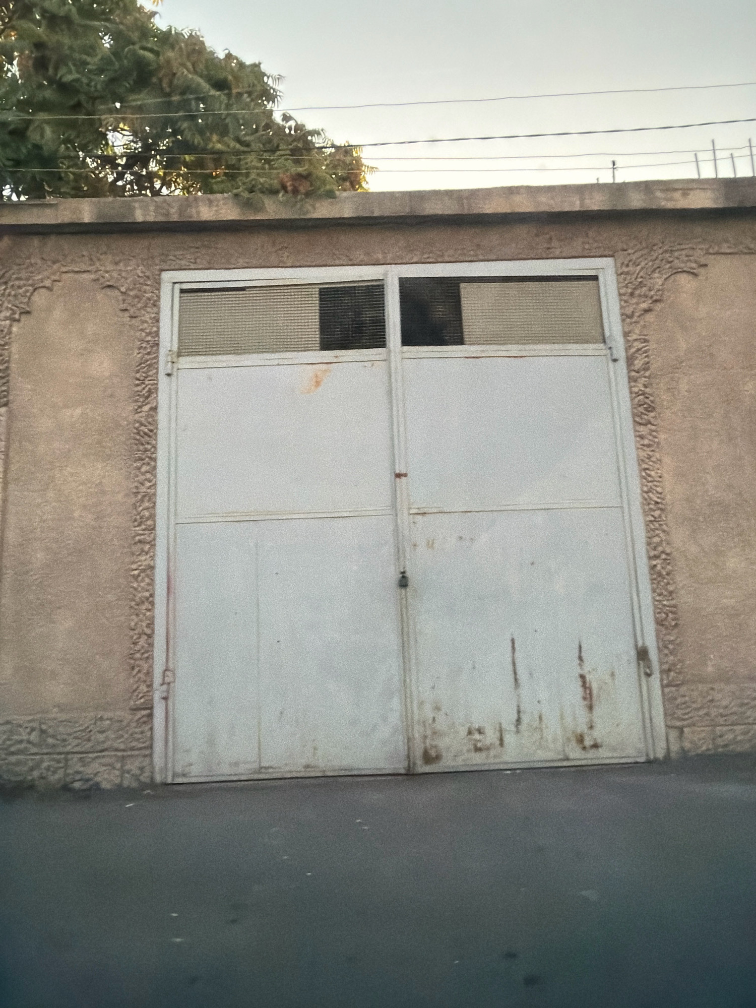 The image depicts a double metal gate, painted a light gray color, set into a textured concrete wall. The gate has visible signs of age and wear, including rust stains and scuffs. Above the gate, there are two rectangular panels with a slatted design, and one of these panels is partially obscured by shadow, revealing a dark interior. The wall itself has decorative, embossed patterns along its sides. Above the wall, a line of barbed wire runs horizontally, indicating a security measure. Trees with dense green foliage are visible behind the barbed wire and to the left of the gate. The ground in front of the gate is asphalt. This scene is located on a street in Yerevan, Armenia, and the overall impression is one of a utilitarian entrance, likely for a garage or a private property, showing signs of weathering and the passage of time within the urban environment of Yerevan.