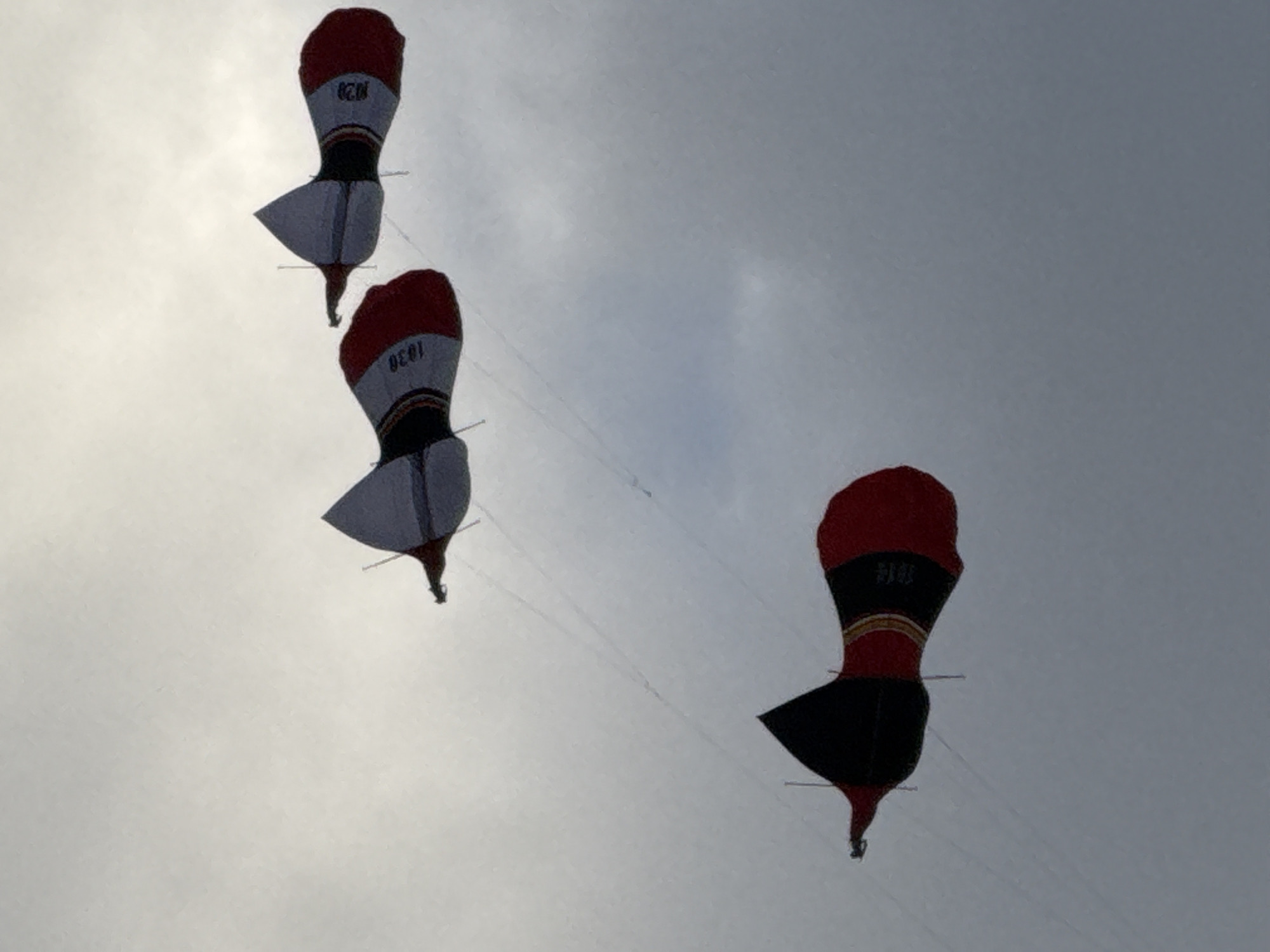 Three kites are flying in a gray, overcast sky. The kites are stylized, resembling rockets or torpedoes, with black, red, and white sections. The top two kites appear identical, featuring the number "1828" printed on their white sections. The bottom kite is similar in shape but has a slightly different color pattern, with broader bands of black and red, and text that is partially obscured. The kites are tethered by thin lines, visible against the sky. The image was taken during daylight, with diffused light suggesting clouds. The location is identified as Denpasar, Indonesia.