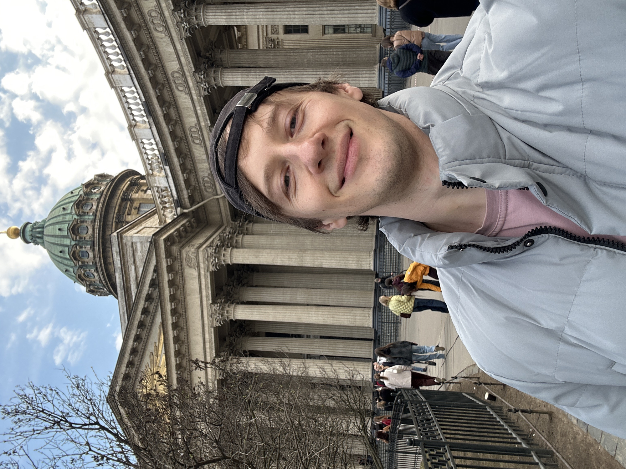 A man in a light gray puffer jacket and a black baseball cap is posing for a selfie in front of Kazan Cathedral in St. Petersburg, Russia. The cathedral's iconic dome and colonnades are visible in the background. There are several other people walking in the area and bare tree branches are in the lower left corner of the frame. The weather appears to be overcast.