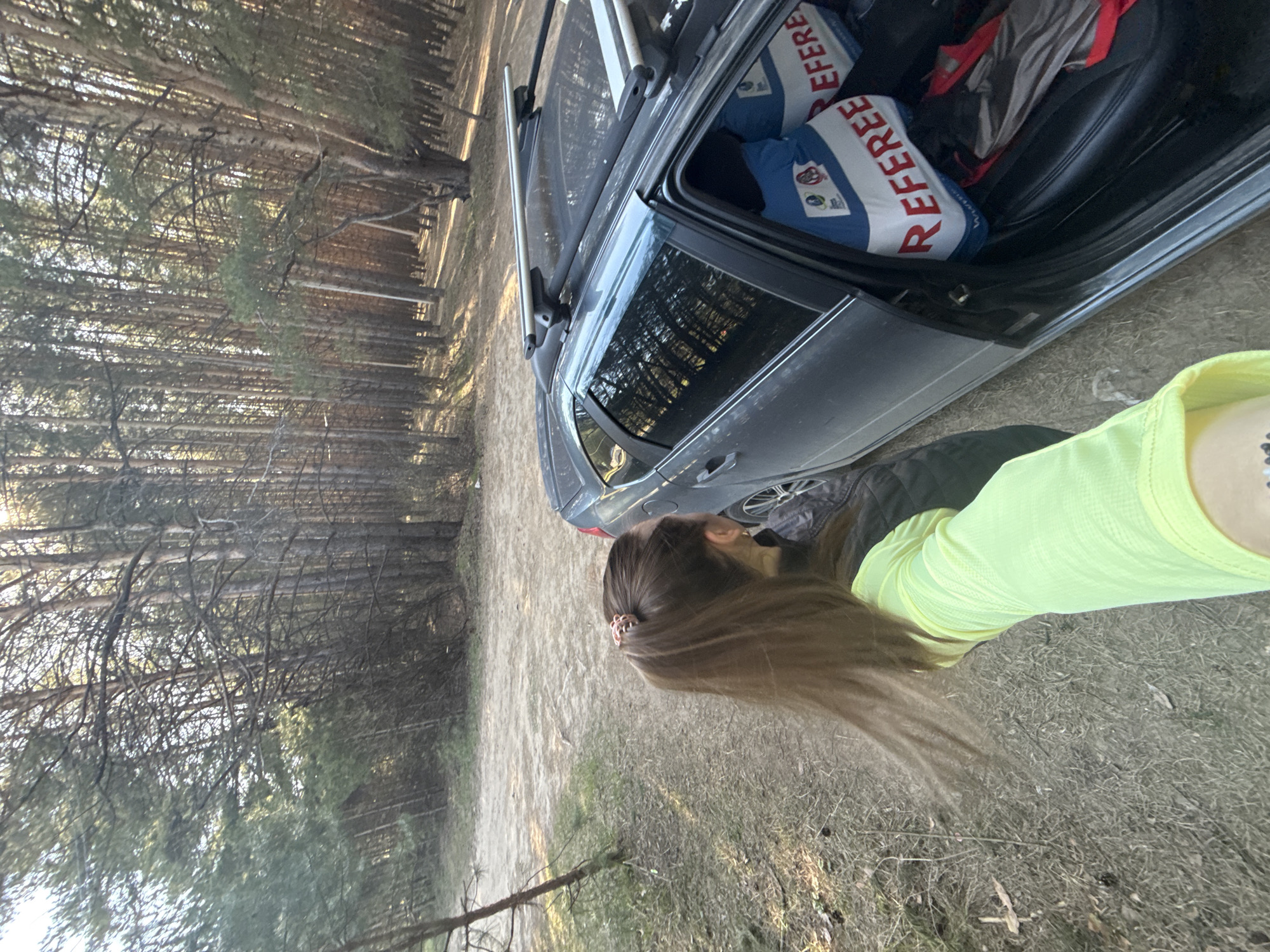 The image, likely taken near the city of Chaikovsky, Russia, shows a scene in a pine forest adjacent to a dirt road. A dark-colored hatchback car with a roof rack has its passenger-side front door open. Inside, several blue and white bags labeled "REFEREE" are visible. A person with long brown hair wearing a bright yellow shirt stands beside the car. The ground is covered in dry grass and pine needles. Tall pine trees dominate the background.
