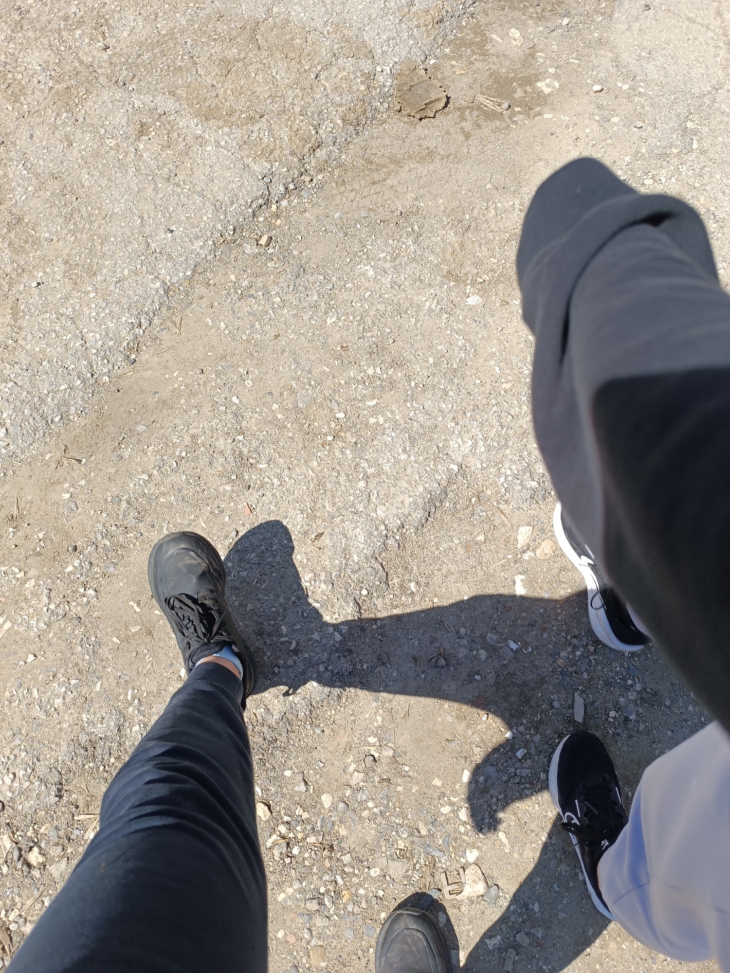 The image depicts two individuals standing on a gravelly, dirt-covered surface, likely outdoors. The person on the left is wearing black shoes and dark pants, while the person on the right is wearing black and white sneakers and light-colored pants. The ground is composed of small rocks and dirt, with some visible cracks and variations in texture. Shadows cast by the individuals and their clothing are also visible. The precise location within Pleven, Bulgaria, cannot be determined from the visual information provided.