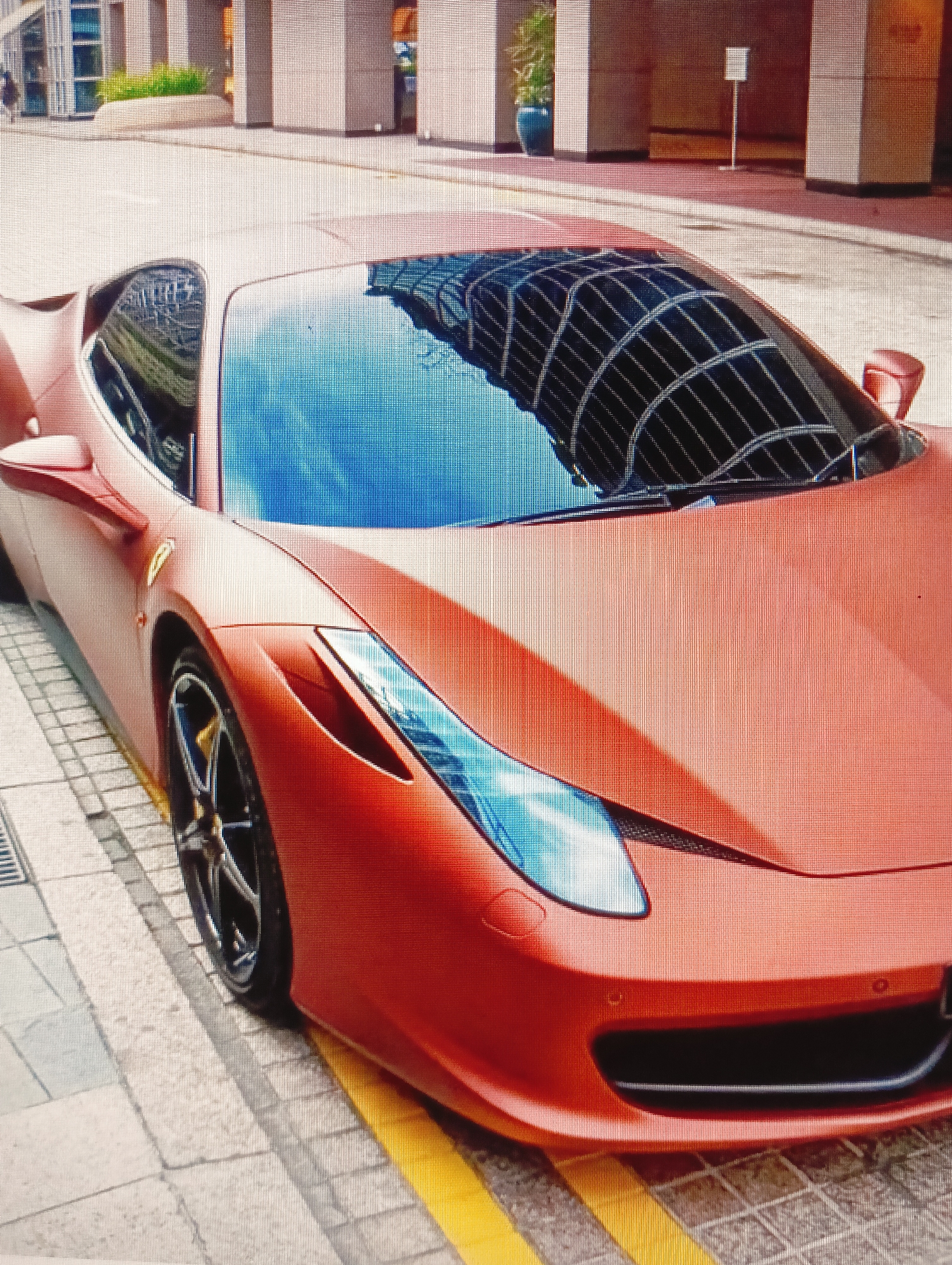 An orange Ferrari sports car is parked on a city street, likely in Sluck, Belarus. The car is positioned at an angle, with its front end slightly turned towards the viewer. The car's matte finish reflects the surrounding environment, including buildings with a grid-like architectural pattern. The street is paved with interlocking stones, and there are yellow lines painted on the road. A drain grate is visible on the left side of the image. The background shows modern buildings and some greenery. No people are clearly visible in the immediate vicinity of the car.