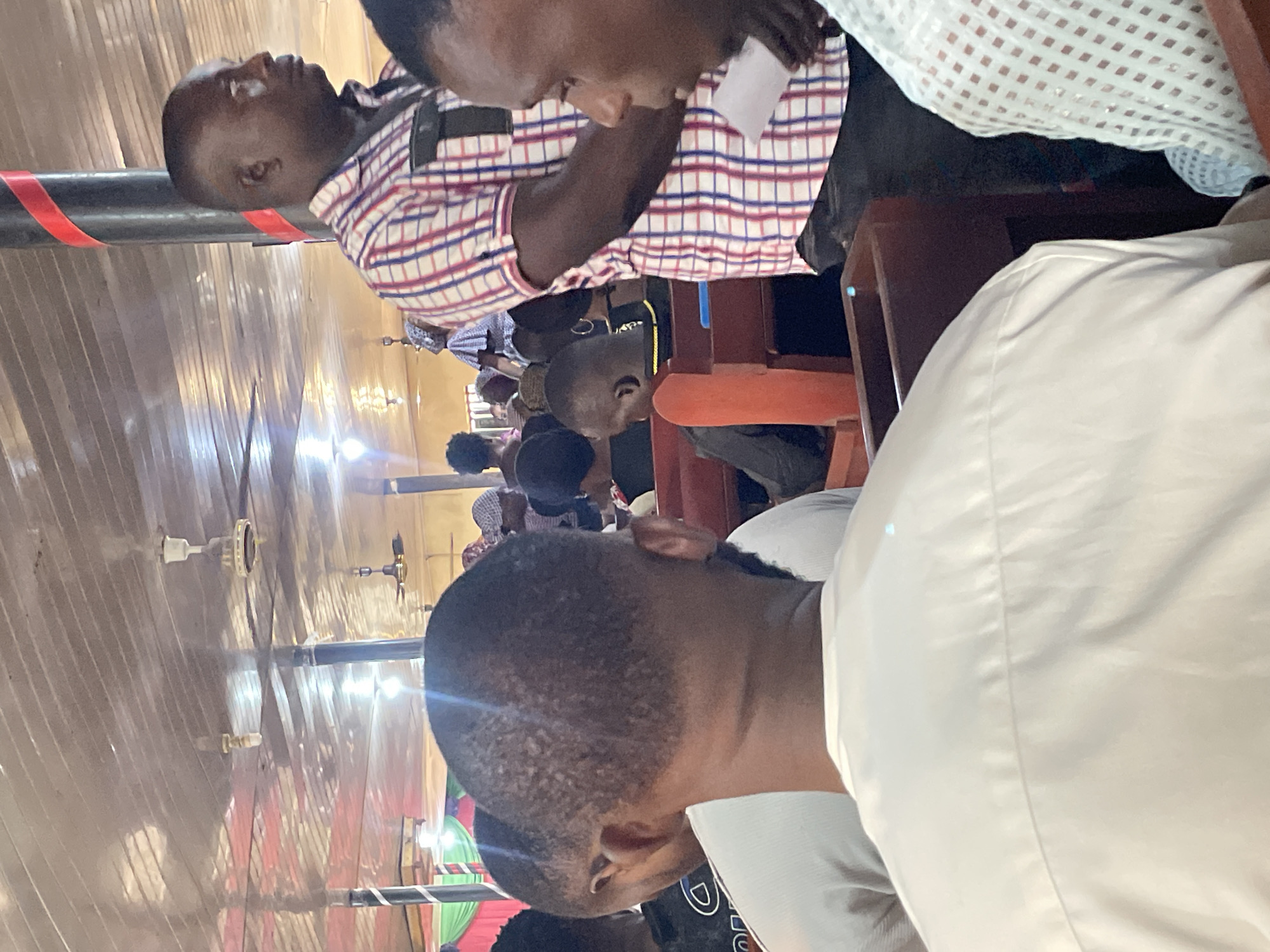 In Abuja, Nigeria, an indoor gathering takes place within a hall, possibly a church or community center, characterized by a sloped, reflective ceiling with visible industrial fans and overhead lighting. The seating arrangement suggests an event or service, with rows of dark wooden benches occupied by attendees.
The foreground is dominated by the backs of several individuals. On the left, a man in a light-colored, possibly white, traditional outfit is seen from the back, with his head and shoulders prominent. To his right, another individual, also seen from behind, wears a black t-shirt with a white circular emblem.
The mid-ground reveals more of the assembly. A man in a red, white, and blue checkered shirt stands, facing to the right, holding a white paper in his hand. His presence suggests he might be a speaker or an usher. Behind him, other attendees are visible, including women with head coverings and children seated in the pews. One person, in a black shirt with a yellow collar, is seated in the middle ground.
The environment is illuminated by artificial lights, and the reflective ceiling creates a polished appearance. The overall impression is of a communal event in a modestly decorated indoor space.