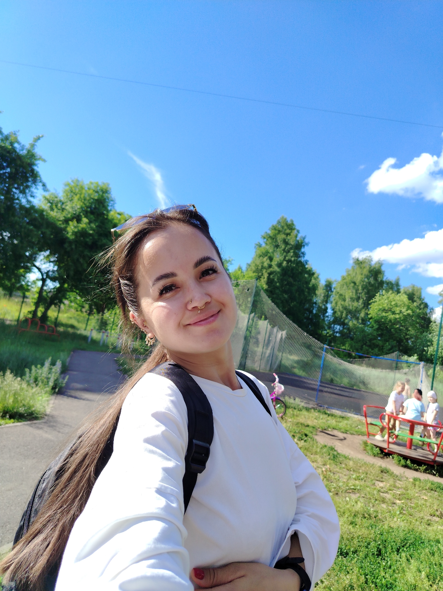 This is a medium shot selfie taken outdoors on a sunny day. The subject is a young woman with long brown hair and a nose ring, wearing a white long-sleeved shirt and a black backpack. She is smiling at the camera, her expression conveying happiness and warmth. In the background, there is a playground with children on a merry-go-round, a net enclosure, and lush green trees under a clear blue sky with scattered white clouds. The scene suggests a casual, recreational outing, likely in a park or outdoor community space in Noviy, Russia. The bright sunlight indicates it is daytime.