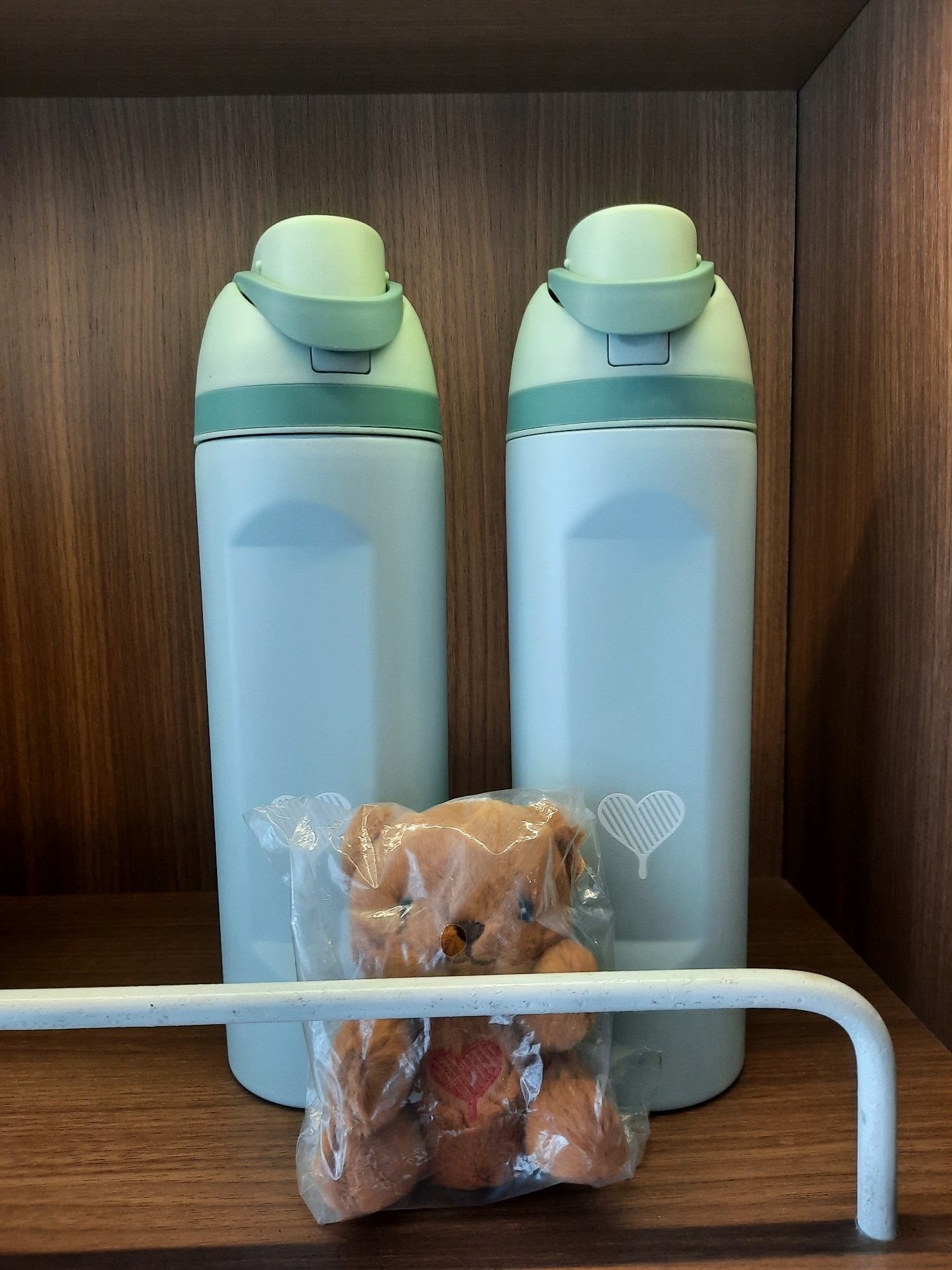 Two light blue thermos flasks with pale green lids stand upright on a wooden shelf. The flasks have a smooth, matte finish. The left flask is slightly obscured by a clear plastic bag containing a small, brown teddy bear. The teddy bear has a stitched red heart on its chest and a white heart design is visible on the right flask. A white metal bar runs horizontally in front of the teddy bear and the lower portion of the flasks. The background consists of vertical wooden planks, indicative of shelving. This scene is located in East Jakarta, Indonesia.