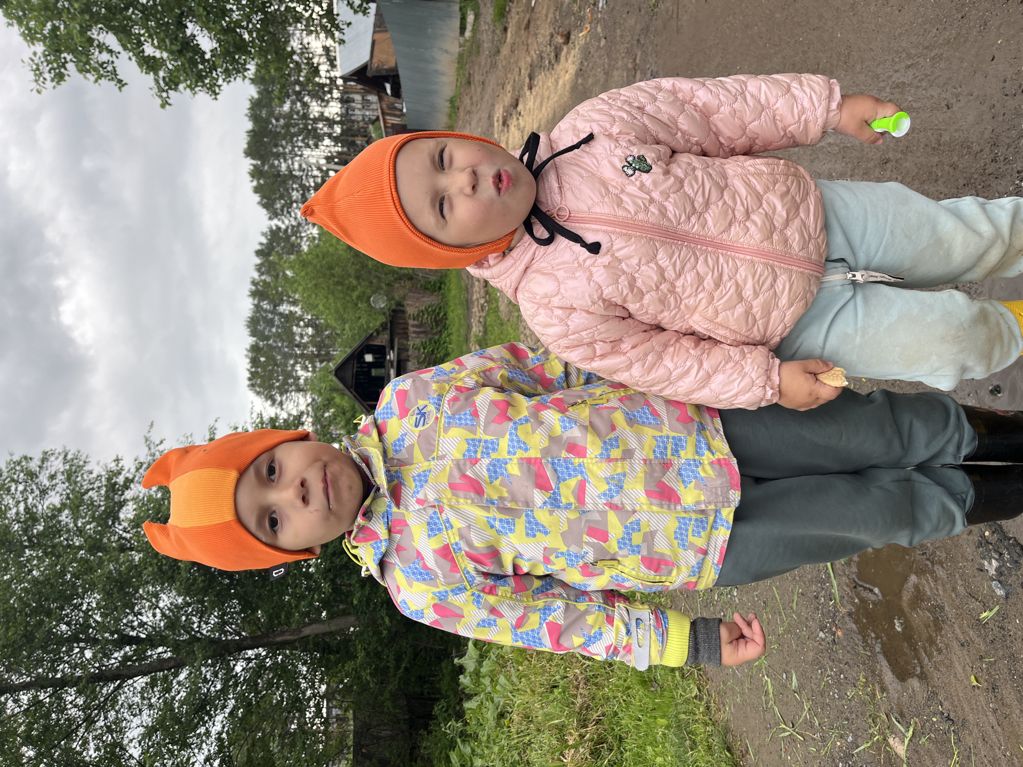 Two young children stand close together outdoors. One child, wearing a bright orange hat with protrusions resembling horns, a multicolored jacket with geometric patterns, and dark-colored pants tucked into black boots, stands facing the camera. The other child, also wearing an orange hat, a pink puffy jacket with a black tie, and light blue pants, stands on top of the first child. The setting appears to be a rural or semi-rural area, with vegetation visible in the foreground and trees and buildings in the background. The ground looks muddy, suggesting recent rain or wet conditions. Location is Chaikovsky, Russia.