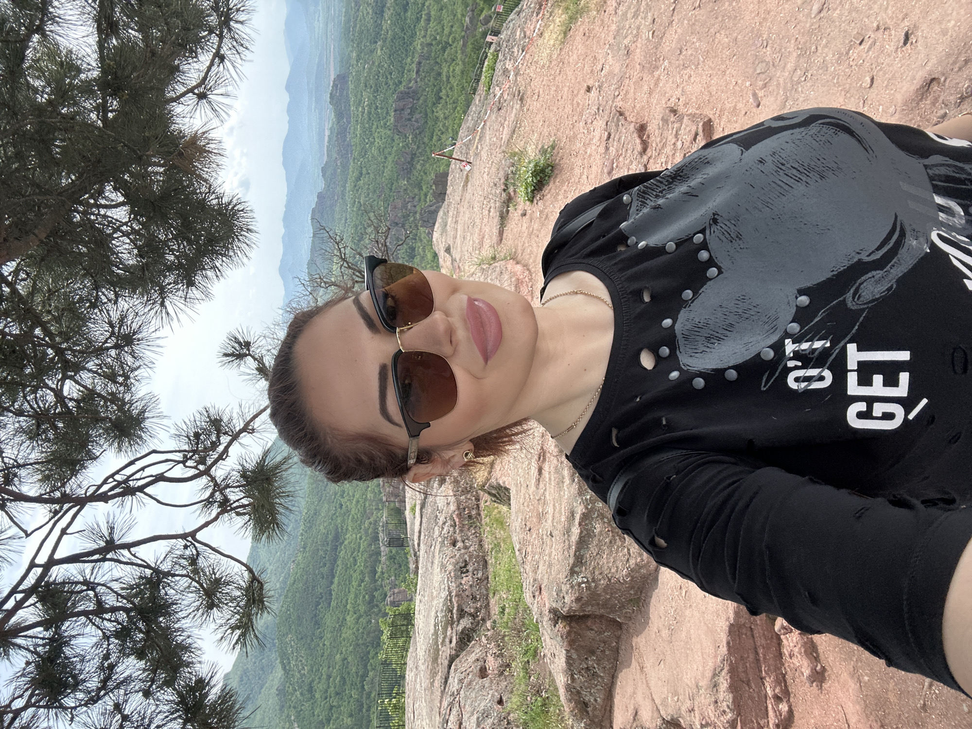 A woman with brown hair pulled back is taking a selfie on a rocky cliff overlooking a landscape of green hills and valleys in Belogradchik, Bulgaria. The woman is wearing sunglasses and a black t-shirt with embellishments and the word "GET" printed on it. A tree branch is visible in the left side of the frame. The ground is rocky and reddish-brown. A railing is visible in the background, suggesting a viewpoint or overlook.