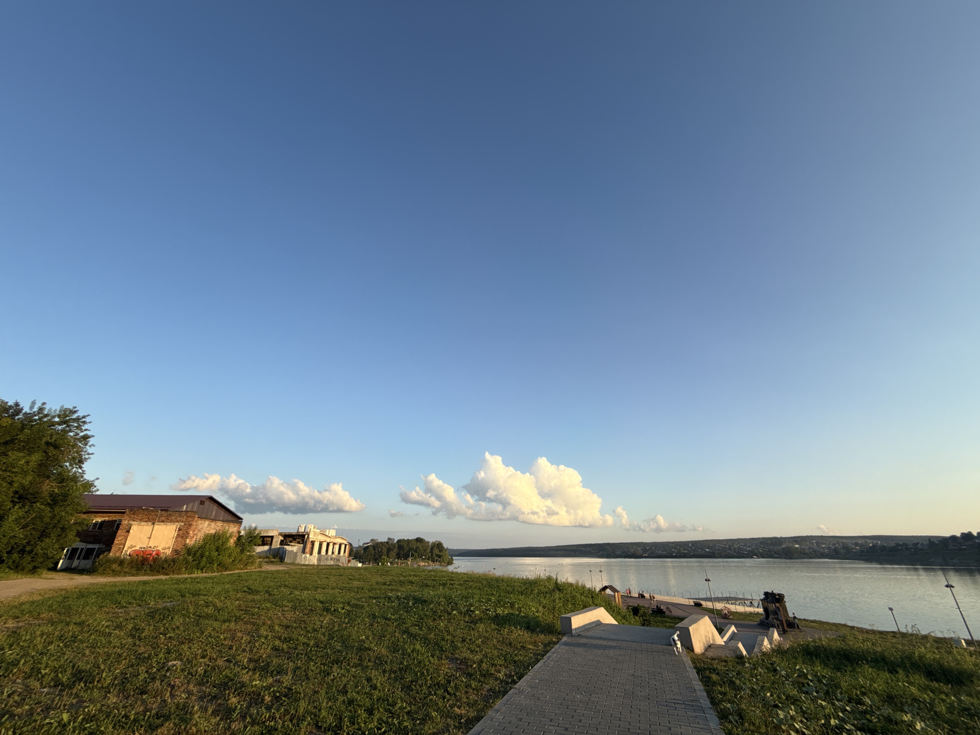 This image captures a serene waterfront scene, likely in the city of Lysva, Russia. The expansive, clear blue sky dominates the upper portion of the frame, with a few scattered white clouds adding texture to the horizon. Below the sky, a wide, calm river reflects the soft light of what appears to be late afternoon or early evening.
On the left side of the image, an old brick building with a brown roof and a large, graffiti-adorned garage door stands amidst overgrown green grass and a dense tree. Further to the right, another building, possibly under construction or renovation, with a white facade and visible window frames, is situated on a slight incline.
A paved walkway, made of grey interlocking bricks, leads diagonally from the foreground into the middle ground. Along the riverbank, decorative elements and what appear to be historical artifacts, possibly artillery pieces, are visible. Several lampposts are strategically placed along the waterfront. A gentle slope covered in lush green grass separates the walkway from the river. In the distance, the opposite bank of the river is lined with trees and appears to be a residential area with scattered houses. The overall impression is one of a peaceful, undeveloped urban waterfront, bathed in the soft, golden light of the setting sun. There are no visible people in the foreground or mid-ground of the image.