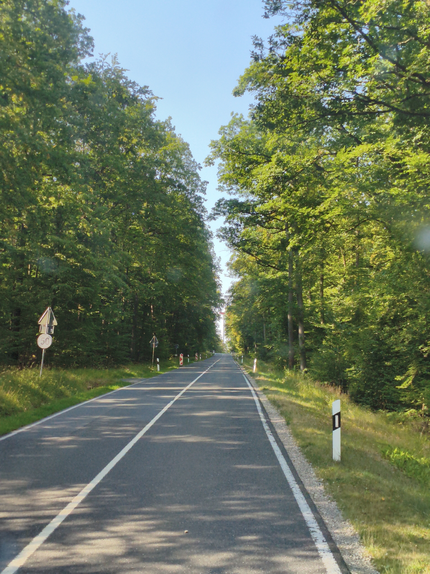 The image shows a road in a forest with trees on both sides of the road. The road is paved and has a white line down the middle. There are traffic signs on the side of the road. The sky is clear and blue. The image appears to be taken from a car driving down the road. The location is the B22 road near Burgwindheim, Germany.
