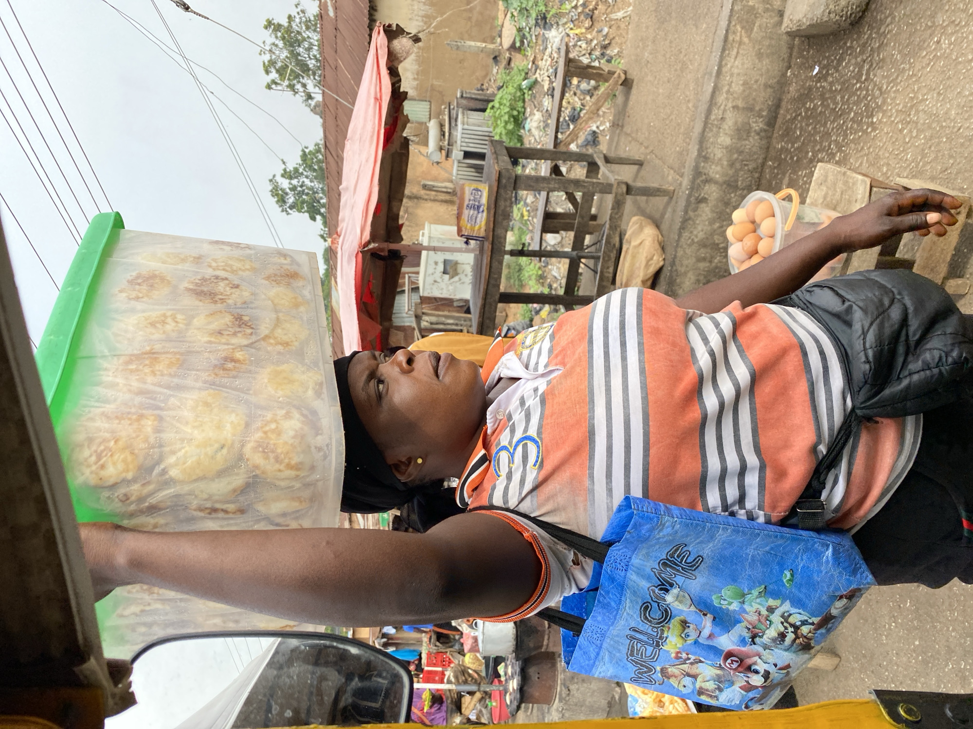 A woman stands near an unidentified mode of transportation in Bukuru, Nigeria. She wears an orange and white striped shirt, a black head covering, and carries a blue shopping bag. A large, clear plastic container, likely containing food items, rests on her outstretched arm. Nearby, a bucket of eggs sits on the ground. The background shows a cluttered urban environment with stalls, buildings, and a small amount of trash visible.
