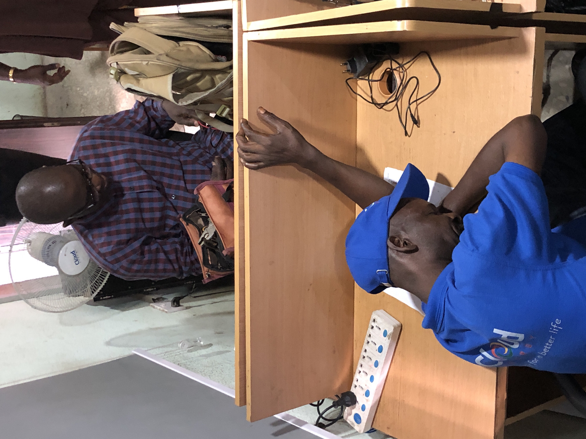 In a dimly lit interior space, likely an office or study area in Ikeja, Nigeria, two Black men are present. In the foreground, a man wearing a blue cap and blue shirt with the text "cloud for better life" printed on it is slumped over a wooden desk, appearing to be asleep or resting his head on his arm. His arm is extended across the desk, and his hand is resting on the edge of a cubicle divider. On the desk in front of him is a white power strip with multiple outlets and a black cord plugged into it. A power adapter with cords is also visible on the desk further back. In the background, another Black man wearing glasses and a plaid shirt is seated, looking down and engaged with a light brown bag and some papers or objects on the desk in front of him. Behind him, a white fan with the logo "Cloud" is mounted on the wall. The environment appears functional, with wooden desks and cubicle dividers suggesting a shared workspace.