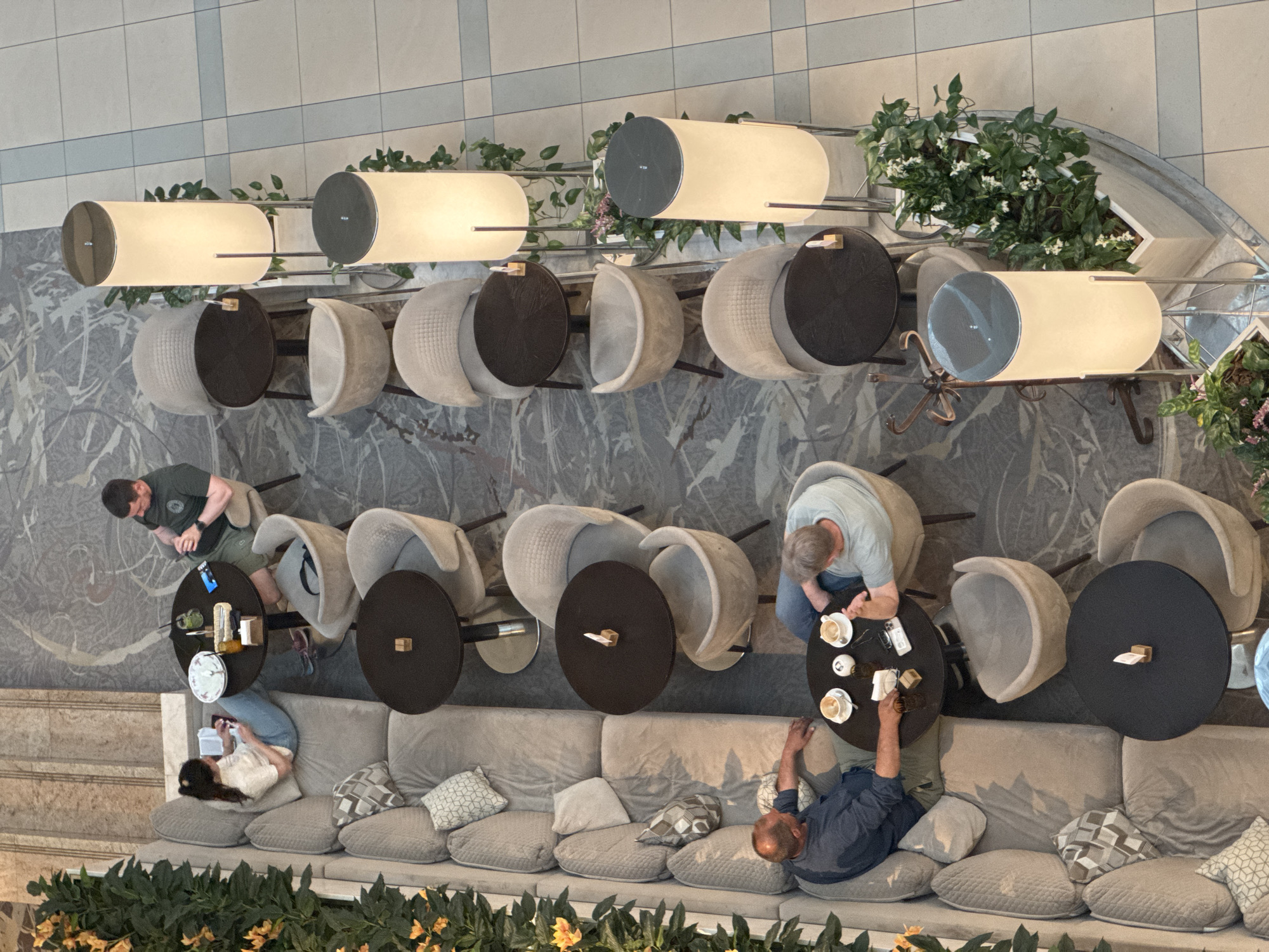 The image shows an overhead view of a seating area in what appears to be a public space, possibly in Moscow, Russia, given the architectural style. The area features a long, cushioned sofa with several decorative pillows. Three people are seated on the sofa: one is lying down and using a phone, and two others appear to be seated at a table, conversing. There are also two other individuals sitting at nearby tables. All of the tables have chairs surrounding them. The area is decorated with greenery, including potted plants and vines. Above the tables and seating area is a row of cylindrical lamps. The flooring consists of light-colored tiles with a subtle pattern. The walls are decorated with marble-like patterns. The overall scene suggests a casual meeting or relaxation area in a public space.