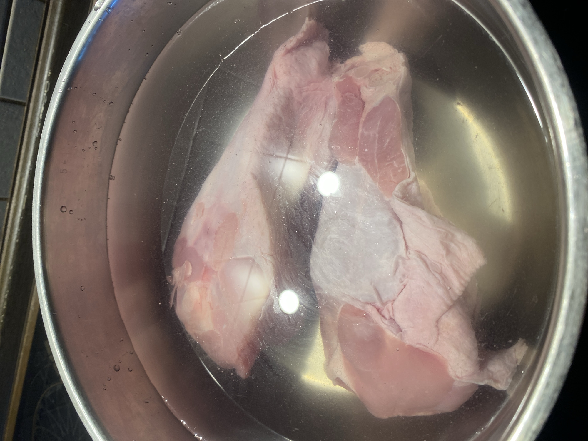 The image shows two raw turkey legs submerged in water in a stainless steel pot. The pot is on a dark surface, possibly a stovetop, with a glimpse of a tiled backsplash in the upper portion of the frame. The water is slightly cloudy, and there are small bubbles on the surface and along the rim of the pot. The turkey legs are light pink and appear to be skin-on. There is a bright reflection on the surface of the water, obscuring some of the details of the meat. No people or specific location in Saint Petersburg are visible.
