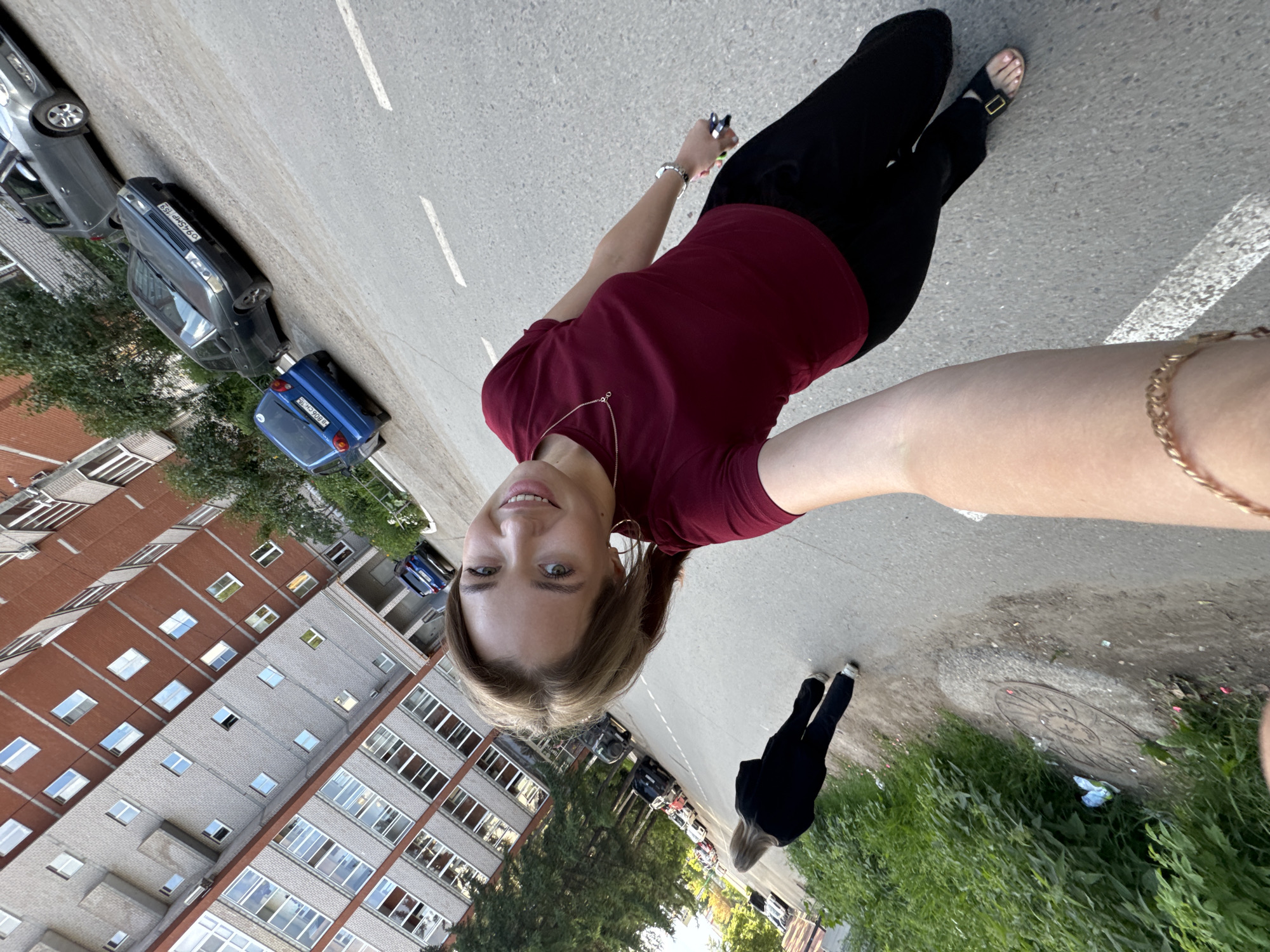 An exterior, eye-level, full-body selfie is taken in Chaikovsky, Russia. The image is dominated by a young woman wearing a maroon t-shirt and black pants, smiling at the camera. She has blonde hair pulled back and is wearing a necklace and bracelet. She is holding a phone in her right hand, and her left arm is extended out of frame. Behind her, a second woman in a black outfit is walking away from the camera, facing towards the right of the frame. The background shows a typical Soviet-era apartment building with grey and red brick facades, numerous windows, and trees lining the street. Several cars are parked along the side of the road. The road surface is asphalt with white lane markings. A patch of tall green grass and some debris are visible on the left side of the image, next to a circular metal manhole cover. The overall scene suggests an urban residential area.