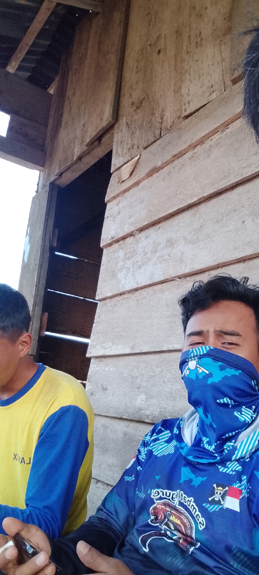 In Padang Dalom, Indonesia, two individuals are depicted in an outdoor setting, possibly under a rustic wooden structure with a corrugated metal roof. The individual on the left wears a yellow long-sleeved shirt with blue accents and a partially visible logo or text. The individual on the right is partially obscured by a blue bandana covering the lower half of their face, which features a pattern of light blue shapes resembling continents. They are wearing a blue long-sleeved shirt with a colorful graphic of a fish and other designs, including what appears to be a Jolly Roger flag and an Indonesian flag. The background consists of weathered wooden planks arranged horizontally, suggesting a simple, possibly unfinished structure. The lighting indicates it is daytime.