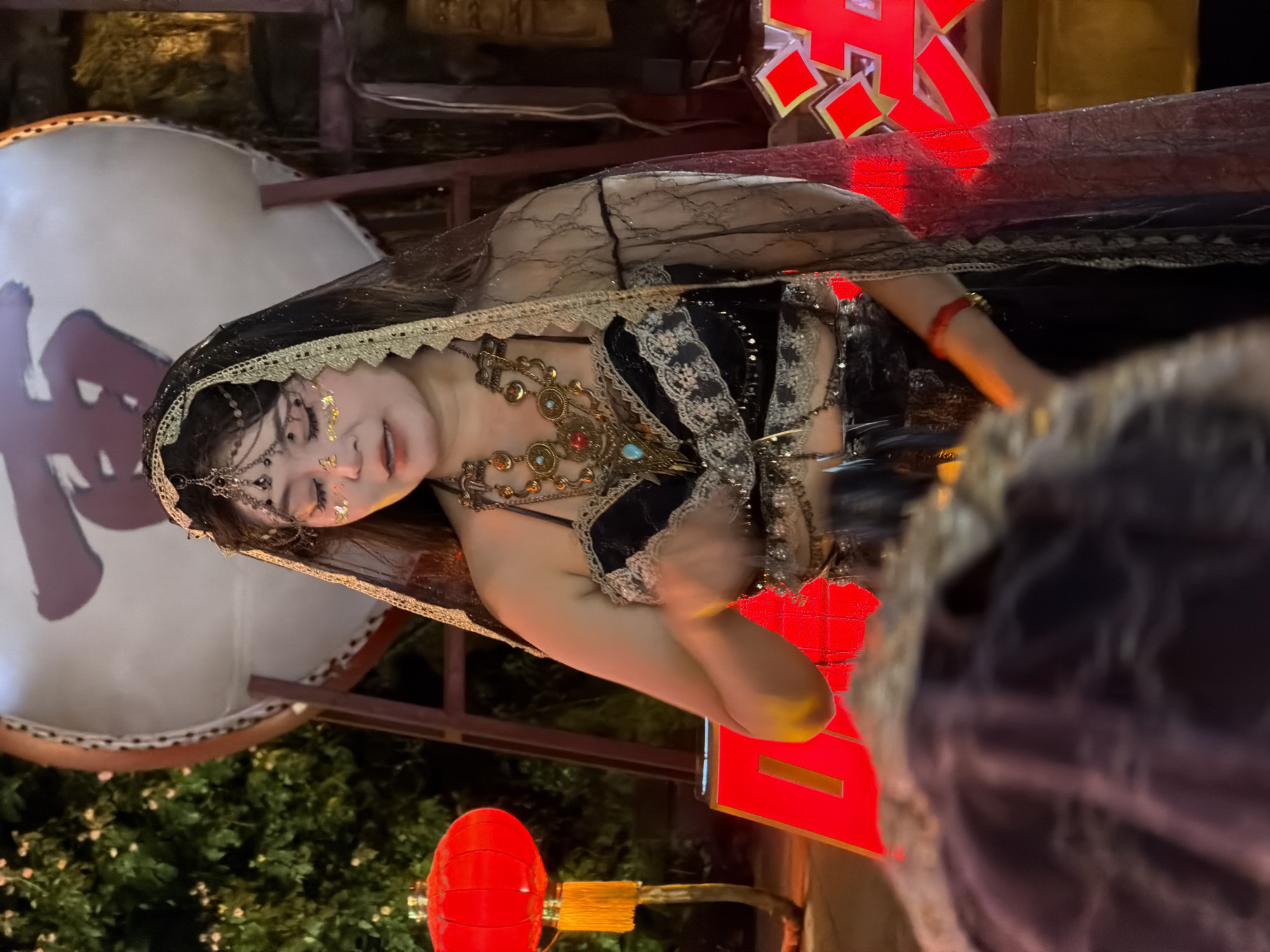 A woman in a black, sequined, belly-baring costume adorned with lace and ornate, gold-toned jewelry, including a multi-layered necklace and a headpiece with dangling beads, stands in front of a large, white drum with a red Chinese character on it. She wears a sheer, black veil over her head and shoulders, edged with gold lace. Her eyes are closed, and glittery makeup with gold flakes is applied to her eyelids and cheeks. Her right hand is raised and blurred with motion. To her left, a red Chinese lantern with a tassel hangs from a branch. In the background, a red structure, possibly a ladder, is visible. To the right, a red sign with glowing geometric shapes is partially obscured by the woman's veil and arm. The overall scene is dimly lit, suggesting it is nighttime, with the red and gold accents creating a festive or theatrical atmosphere. The presence of the drum and Chinese character suggests a setting in China, possibly Yuzhong District.