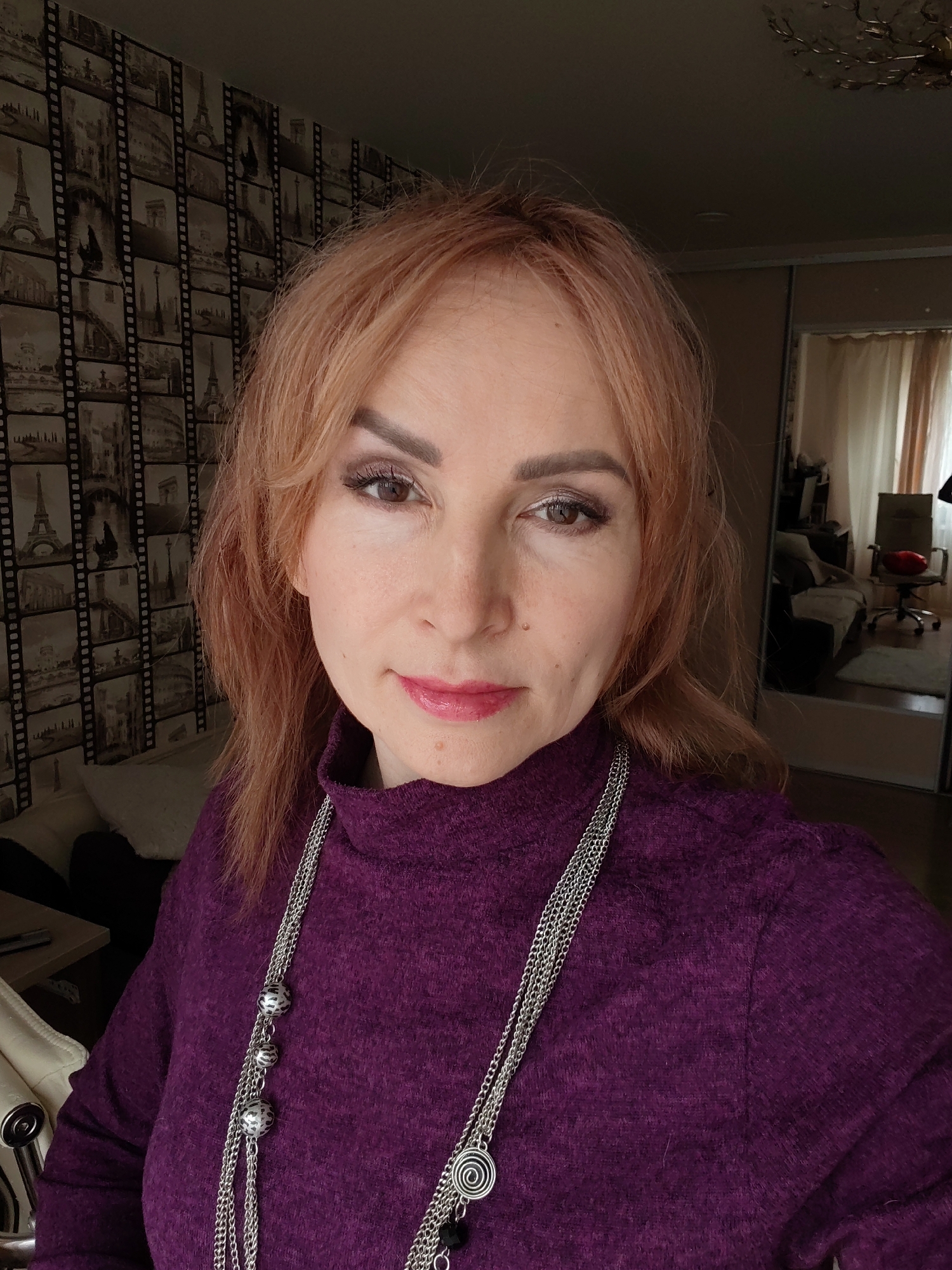 A middle-aged woman poses for a close-up portrait indoors. She has fair skin, light reddish-blonde hair, and is wearing a dark purple turtleneck sweater and a long, multi-strand silver necklace with decorative beads and pendants. She has light makeup, including eye shadow and lipstick.
Behind her, the wallpaper is a pattern of film strips depicting scenes of famous landmarks such as the Eiffel Tower and the Arc de Triomphe. A mirror in the background shows a glimpse of another room with a desk chair and a couch.
There is no definitive information in the image to confirm its location in Chaikovsky, Russia. This would require external information not visible in the image.