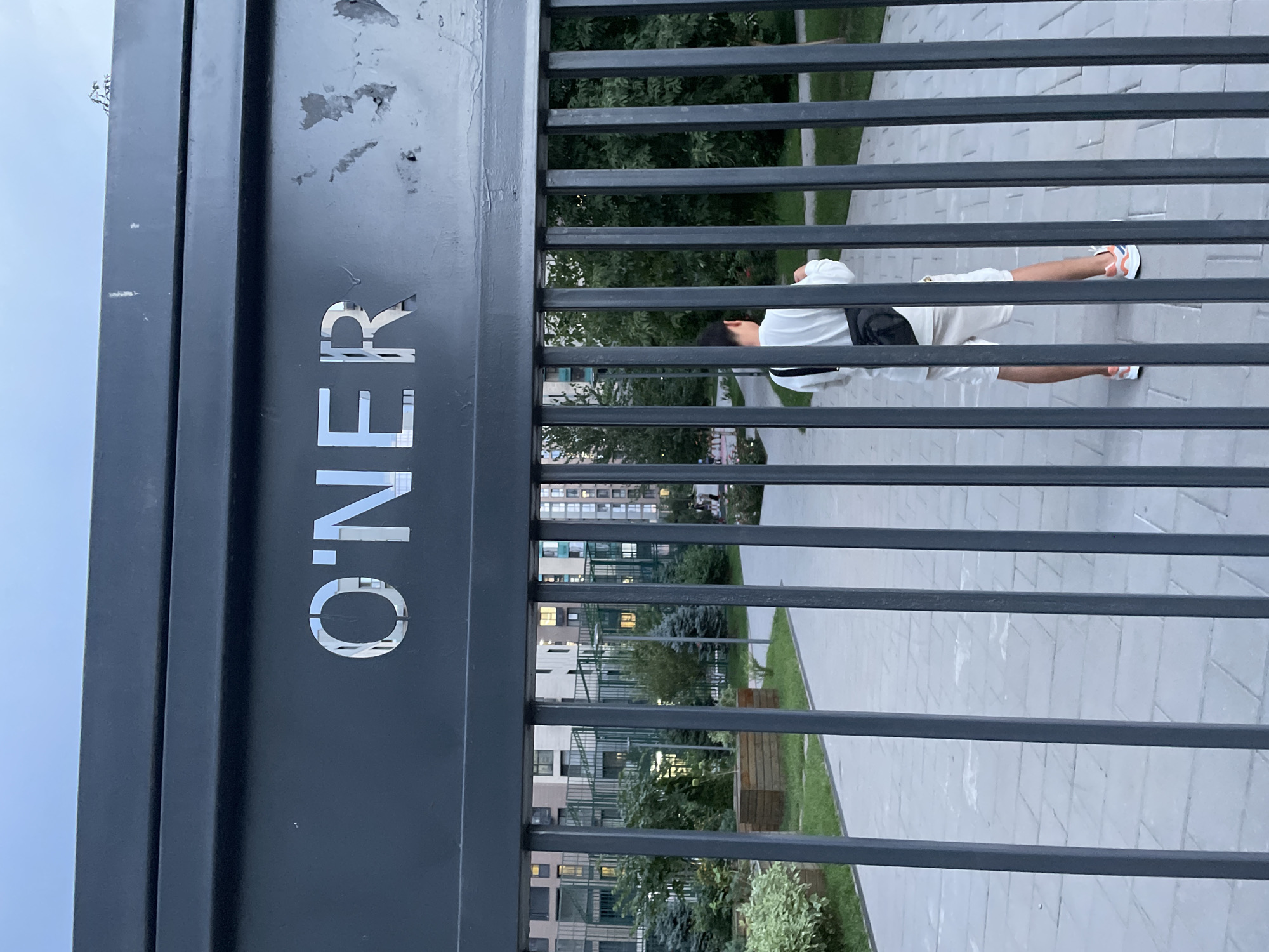 A person wearing a white long-sleeved shirt, beige shorts, and colorful athletic shoes stands facing away from the camera, looking into a courtyard through a black metal gate. A black crossbody bag is slung over their shoulder. The gate has raised, silver block letters spelling "O'NER" on the horizontal bar at the top. Behind the gate, a paved walkway leads through a landscaped courtyard with green trees, bushes, and wooden planters. In the background, modern apartment buildings with lit windows are visible. The scene appears to be in a residential complex. The lighting suggests it is either early morning or late evening. The image does not provide any specific visual cues to identify the location as Almaty, Kazakhstan.