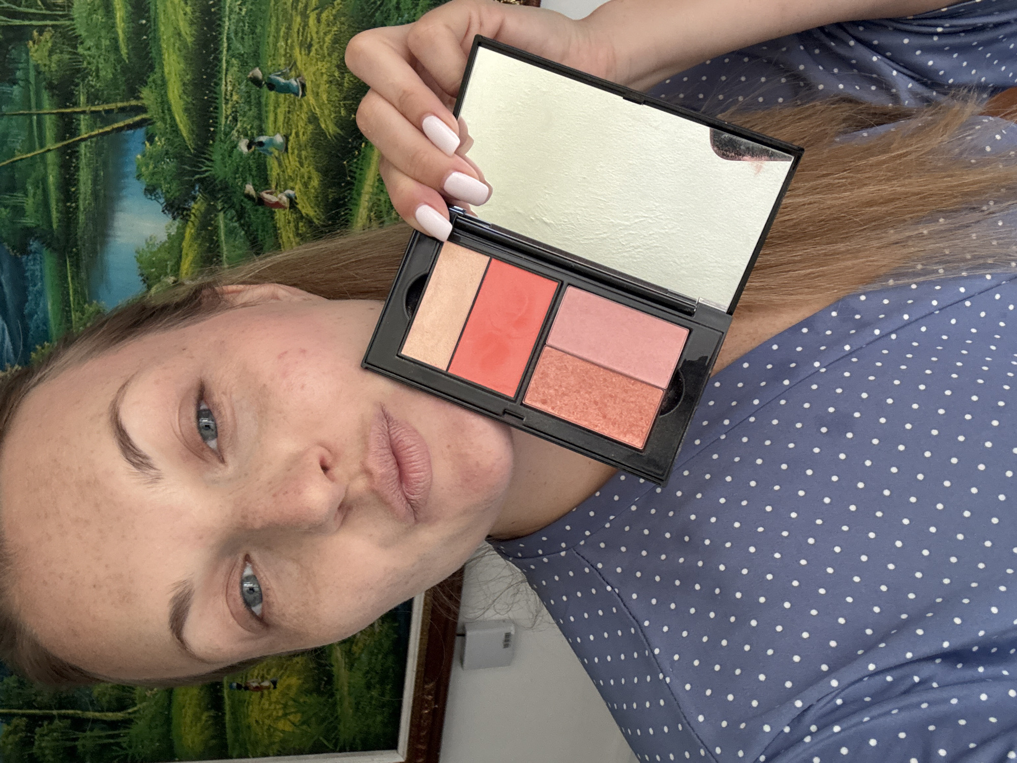 A woman with light skin and freckles holds a makeup compact open in front of her face, her lips pursed. She wears a blue polka-dotted robe. The makeup compact contains four pans of blush and highlighter, and a mirror. Behind her is a painting depicting a landscape with a body of water and three figures. The location is Denpasar, Indonesia.