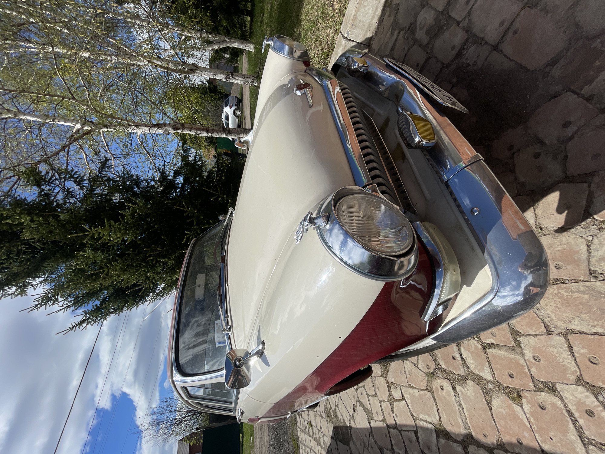 The image shows a two-tone cream and burgundy vintage car parked on a brick-paved surface. The location is Минино, Russia. The car's front end is prominently displayed, with a large, round headlight and chrome bumper visible. In the background, birch trees, a dark green coniferous tree, and a glimpse of a modern white car can be seen. The sky is blue with scattered clouds. Power lines are also visible in the background.