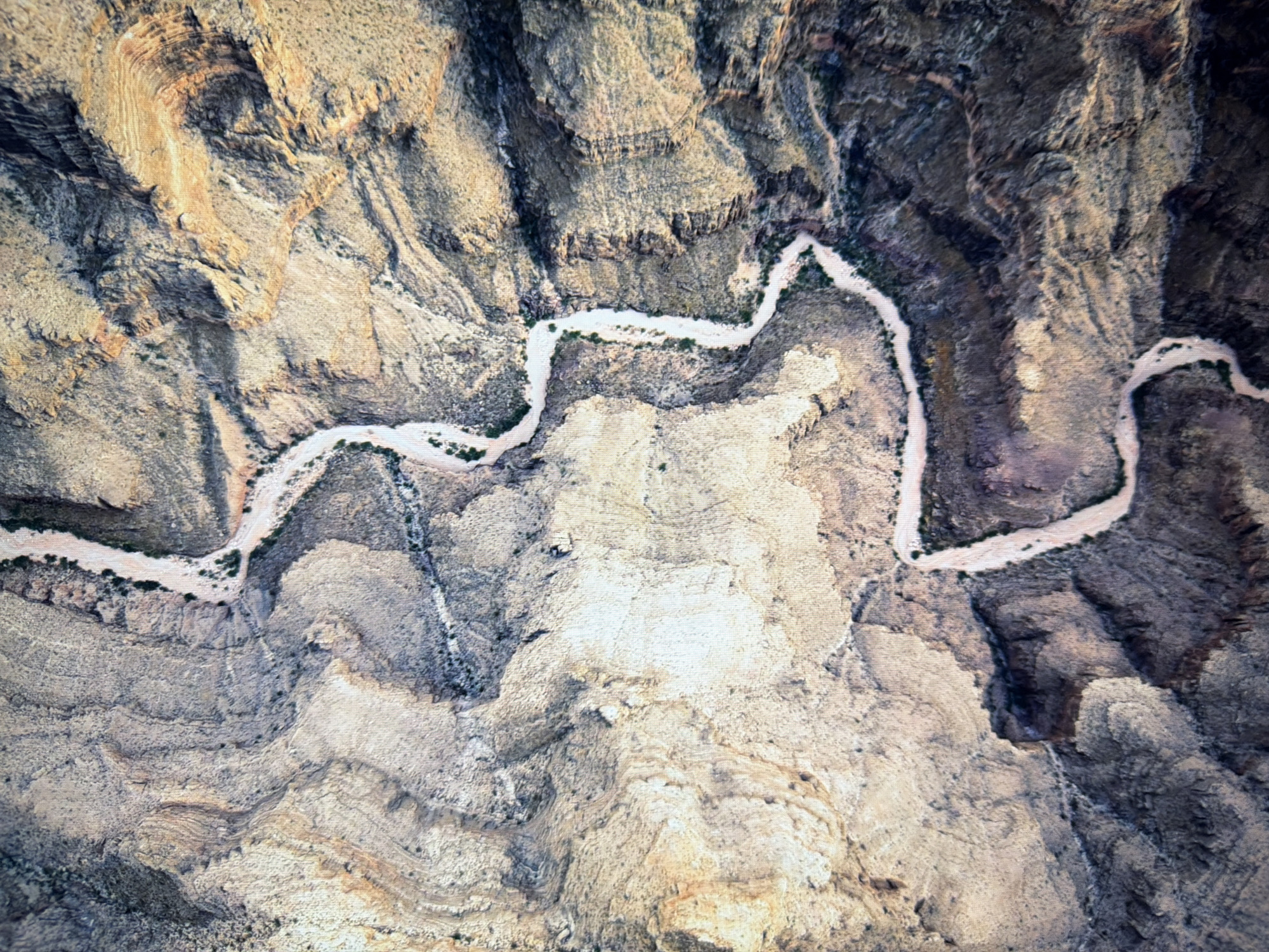 This is an aerial, top-down view of a dry, winding riverbed flowing through a rocky, mountainous landscape. The terrain is characterized by steep, eroded slopes with varying shades of brown, tan, and gray rock formations. Sparse vegetation, appearing as dark green or brown patches, is visible along the edges of the riverbed and on some of the rocky outcrops. The riverbed itself is a lighter, sandy color and appears to be dry, with no visible water. The image does not depict any urban environment, people, or specific geographical landmarks, and therefore cannot be associated with the city of Kyiv, Ukraine.
